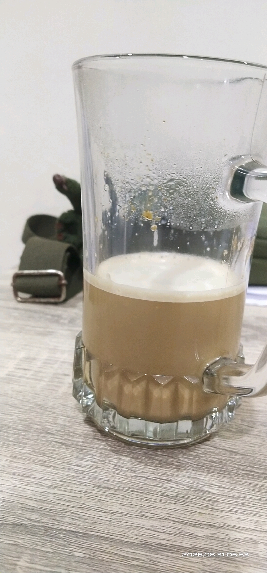A close-up, eye-level shot shows a clear glass mug filled with a frothy, light brown beverage, likely coffee or tea, resting on a light-colored, wood-grain textured surface. The glass is partially filled, with condensation visible on the exterior, suggesting the beverage is hot. The mug has a thick, patterned base and a handle is visible on the right side. In the background, slightly out of focus, is a green strap with a silver buckle. The environment appears to be indoors, possibly a desk or table setting, with a plain white background. A timestamp in the bottom right corner reads "2026.08.31 05:53". There is no indication of the specific location being Matabu, Indonesia.