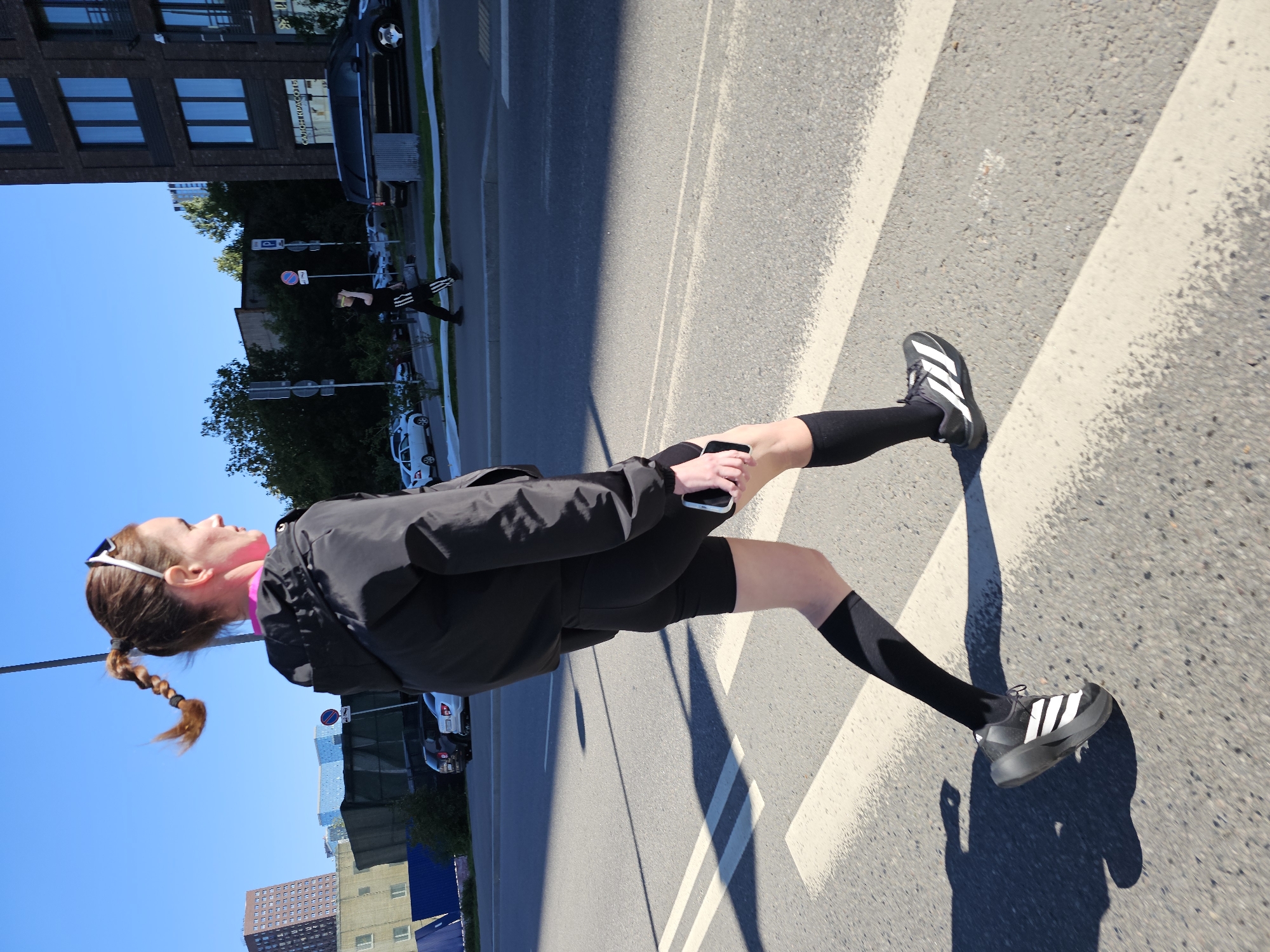 A woman with her hair in a braid is crossing a pedestrian street in Moscow, Russia, on a sunny day. She is wearing a black jacket, black shorts, black socks, and black and white athletic shoes. She is holding a smartphone in her right hand. In the background, there are buildings, trees, and cars. A man is also visible crossing the street in the distance. The scene is a street with a crosswalk.
