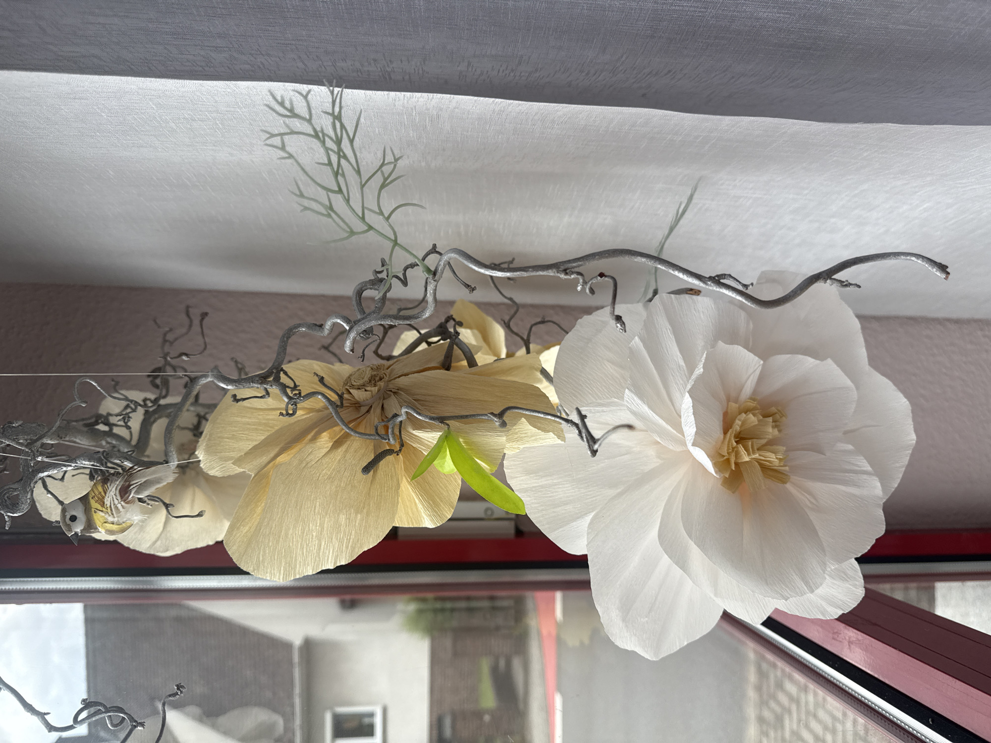 A close-up, indoor shot shows two artificial flowers hanging in front of a window. The flower in the foreground is large and white, with multiple delicate petals layered to create a full blossom. The center of this flower reveals a cluster of smaller, light yellow petals. Behind it, a second, larger flower is visible, made of translucent, pale yellow paper with ruffled petals. A small, realistic-looking artificial bird with brown and yellow markings is perched on a silver, twisted wire structure that hangs with the flowers. A single green leaf is attached to the wire near the yellow flower. The background includes a window with a red frame, through which a street and houses in a suburban setting can be seen, consistent with a location in Heiligenrode, Germany. A white curtain is draped to the right of the flowers, partially obscuring the window view and the wall behind. The scene is lit by natural light.