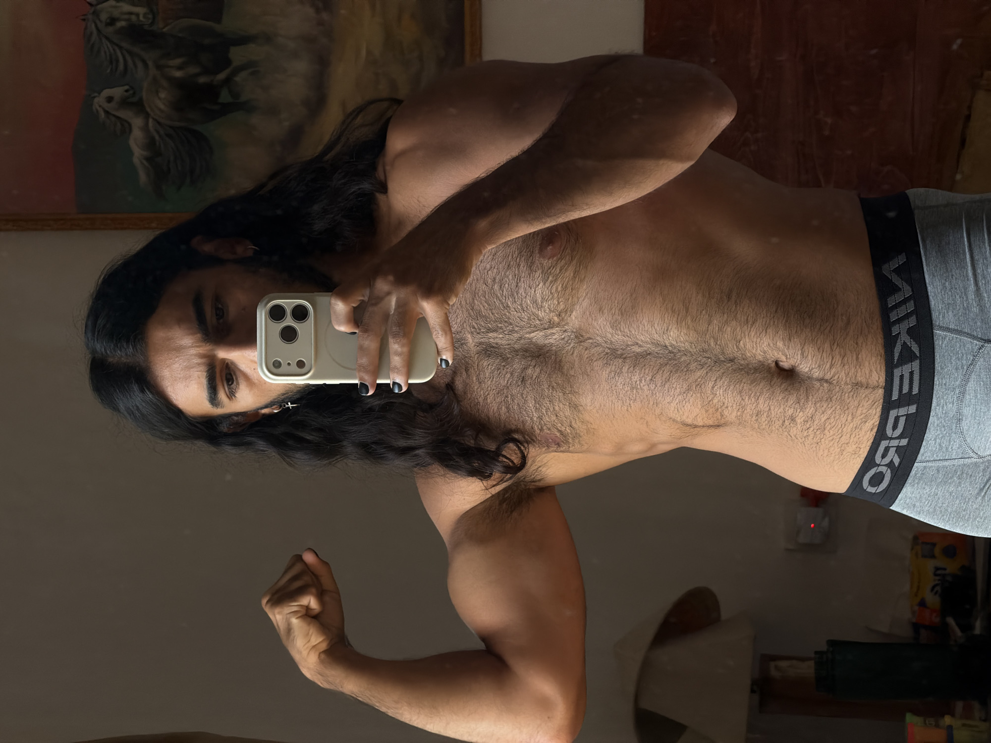 A man with long, dark hair, wearing grey boxer briefs, poses for a mirror selfie in a room. He is flexing his bicep with his left arm, and holding a smartphone with his right hand. His chest and abdomen are visible and covered in dark hair. In the background, a painting of horses and a lamp are visible. The scene appears to be indoors, possibly in a bedroom, with a plain wall and wooden accents. The lighting is dim, suggesting it may be daytime with the blinds drawn or evening.