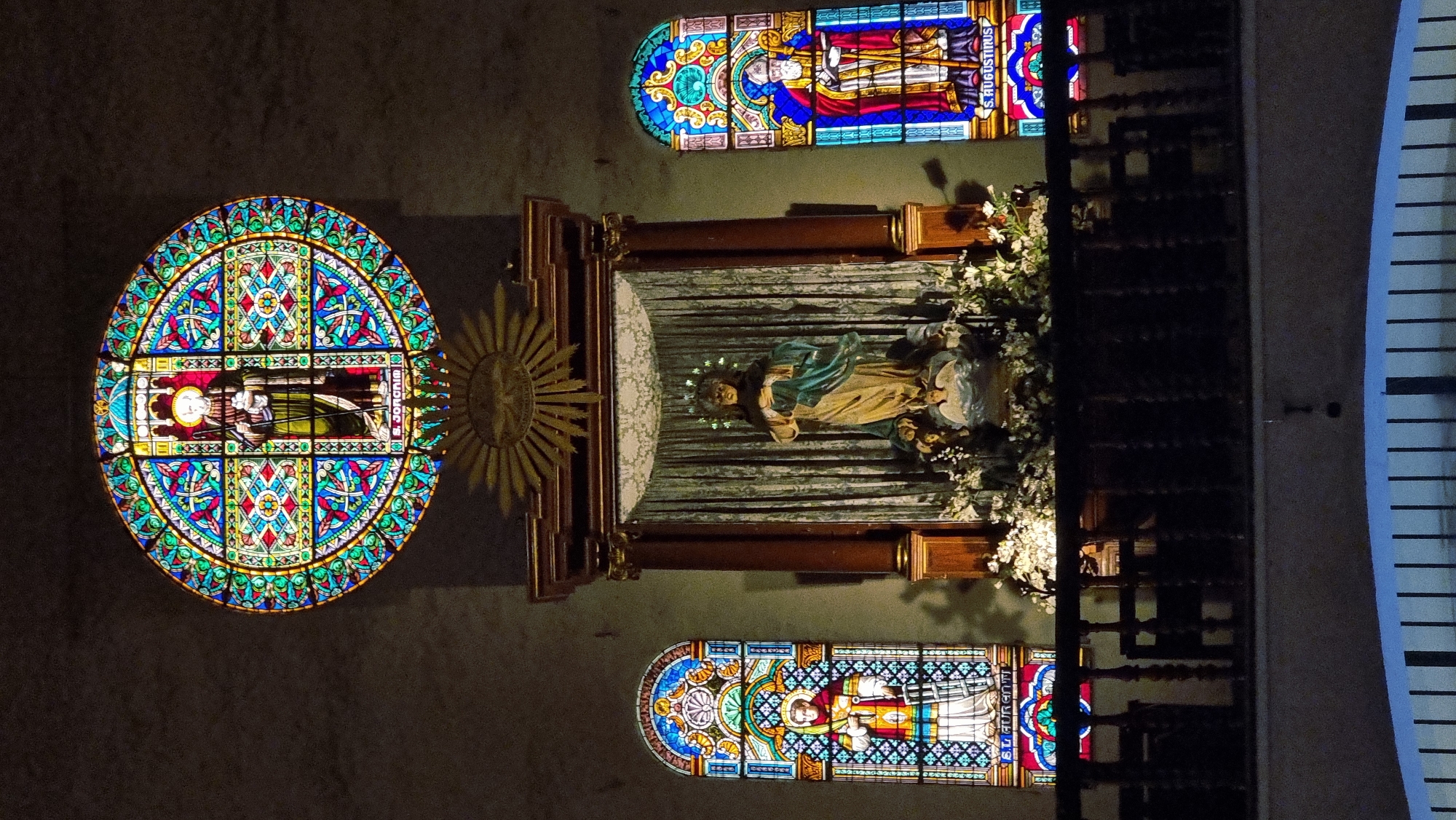 The image shows the interior of a dimly lit church, likely in Cadaqués, Spain. The scene is dominated by religious iconography and architectural elements.
In the center, a statue of a robed figure, possibly a saint or religious icon, is displayed in an alcove. The statue is adorned with a halo and surrounded by a profusion of white flowers. Behind the statue, a draped curtain creates a dramatic backdrop. To the left of the statue, a large, circular stained-glass window features intricate geometric patterns and a central depiction of a person in religious attire. Adjacent to this window is a golden, sunburst-like emblem.
Above and below the central display are two rectangular stained-glass windows. The upper window depicts two figures in religious vestments, with visible text on the right side that reads "S. AUGUSTINUS". The lower window also features a religious figure, though the text is less legible.
The overall atmosphere is solemn and reverent, consistent with a religious setting. The lighting suggests it is either evening or a time when artificial illumination is needed within the church. The architectural style and the presence of stained-glass windows are characteristic of European churches, and the specific mention of "S. AUGUSTINUS" may refer to Saint Augustine, a prominent figure in Christian theology and history.