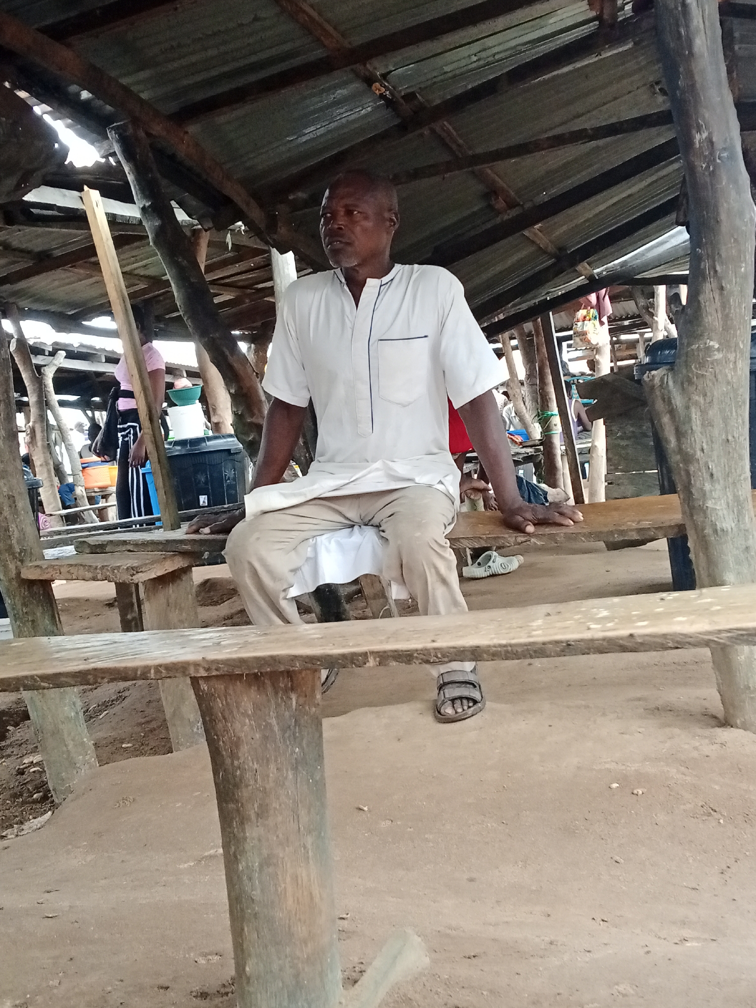 A man is sitting at a wooden table in an outdoor market. The man is wearing a white shirt and beige pants. He is sitting on a wooden bench. The table and benches are made of rough-hewn wood. The market appears to be covered by a corrugated metal roof supported by wooden poles. In the background, other people and market stalls can be seen. The ground is made of dirt.