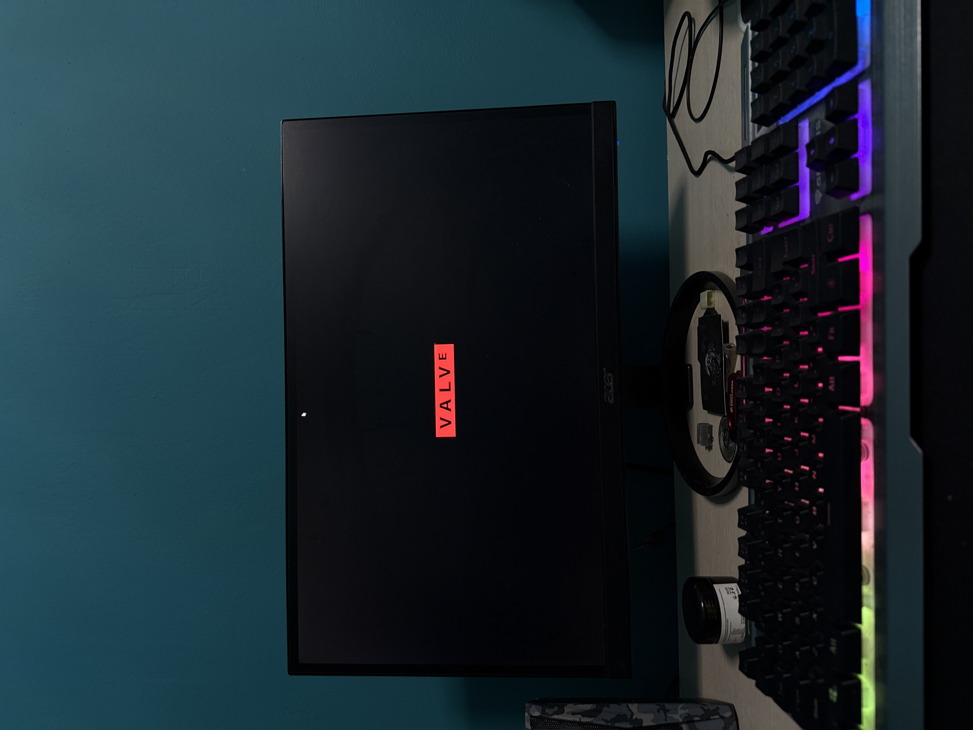 The image shows a computer setup. A black Acer monitor is prominently displayed, showing the Valve logo. To the right of the monitor is a gaming keyboard with RGB lighting. The keyboard is mounted vertically on the wall. A circular decorative item is also present behind the keyboard. A blue wall is visible in the background. No people are visible. The location is unknown.