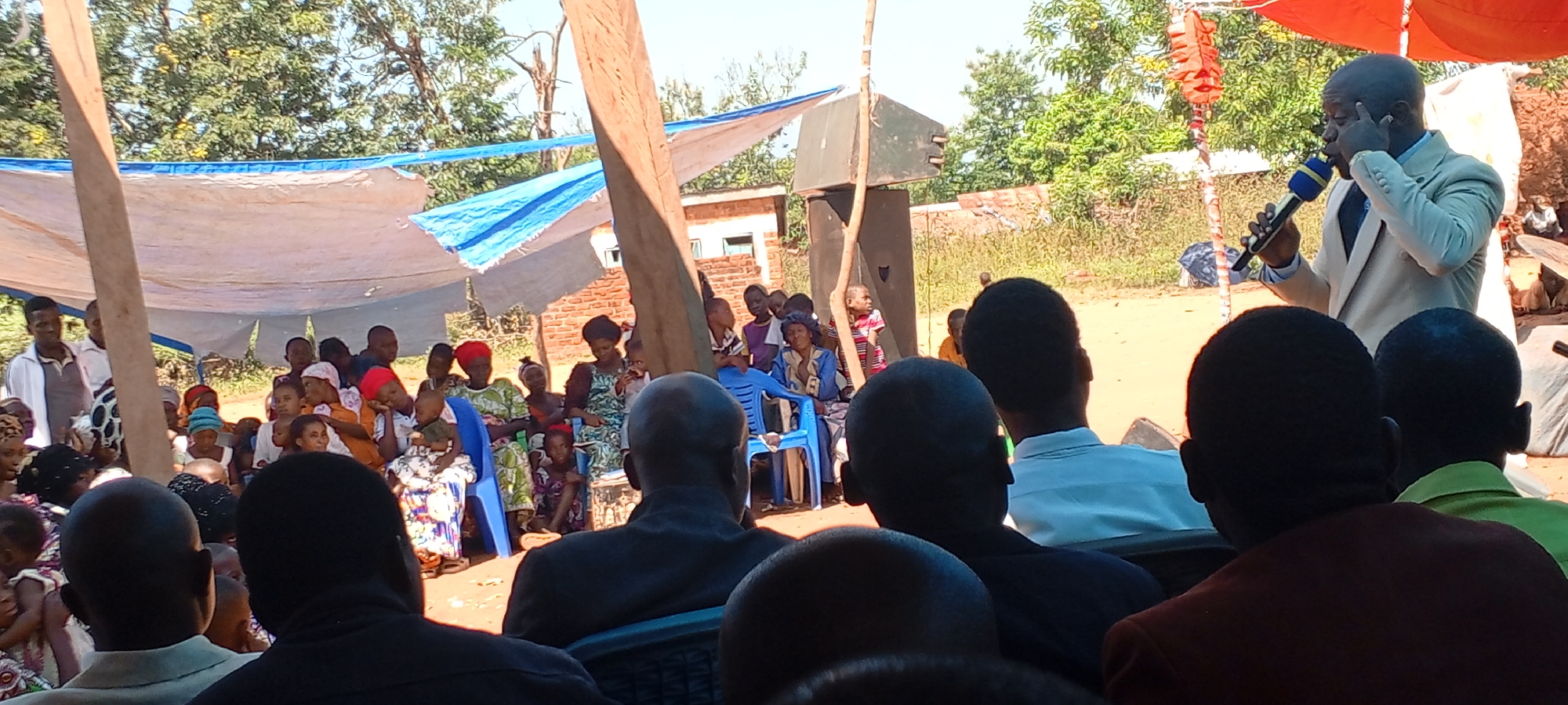 A daytime scene captures a gathering of people outdoors. An individual in a light-colored suit stands at the front, speaking into a microphone. A crowd of people of varied ages and clothing sit on chairs, mostly facing the speaker. The event is taking place under a canopy of white and blue tarps strung between wooden poles. A loudspeaker sits beside a pole, suggesting an outdoor event setup. The background includes trees and foliage. The context suggests an event or meeting, possibly in the city of Kalemie, Democratic Republic of the Congo.