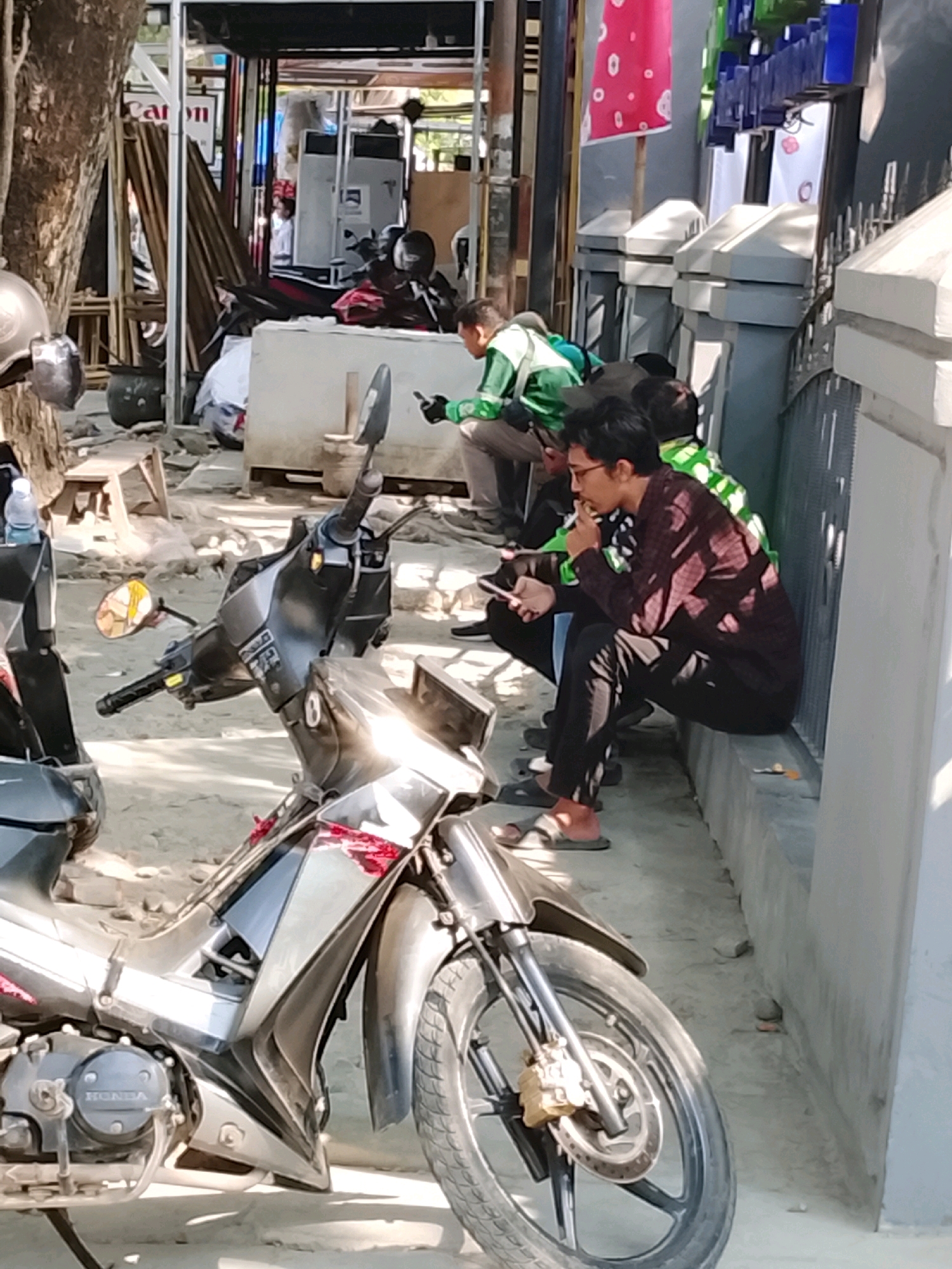 In Slawi, Indonesia, on a sunny day, three men are seen. One man in the foreground, seated on a motorcycle, is wearing a patterned brown long-sleeved shirt and black pants. He holds a mobile phone in his left hand and appears to be smoking. To his right, another man in a green jacket sits on a concrete ledge, also looking at his phone. A third man, also in a green jacket, is partially visible further to the right, also engaged with his phone. The setting appears to be an outdoor area with a concrete wall and fence on the right, and a street or path with motorcycles parked. A tree trunk is visible on the left side of the image. The ground is covered in dirt and gravel.