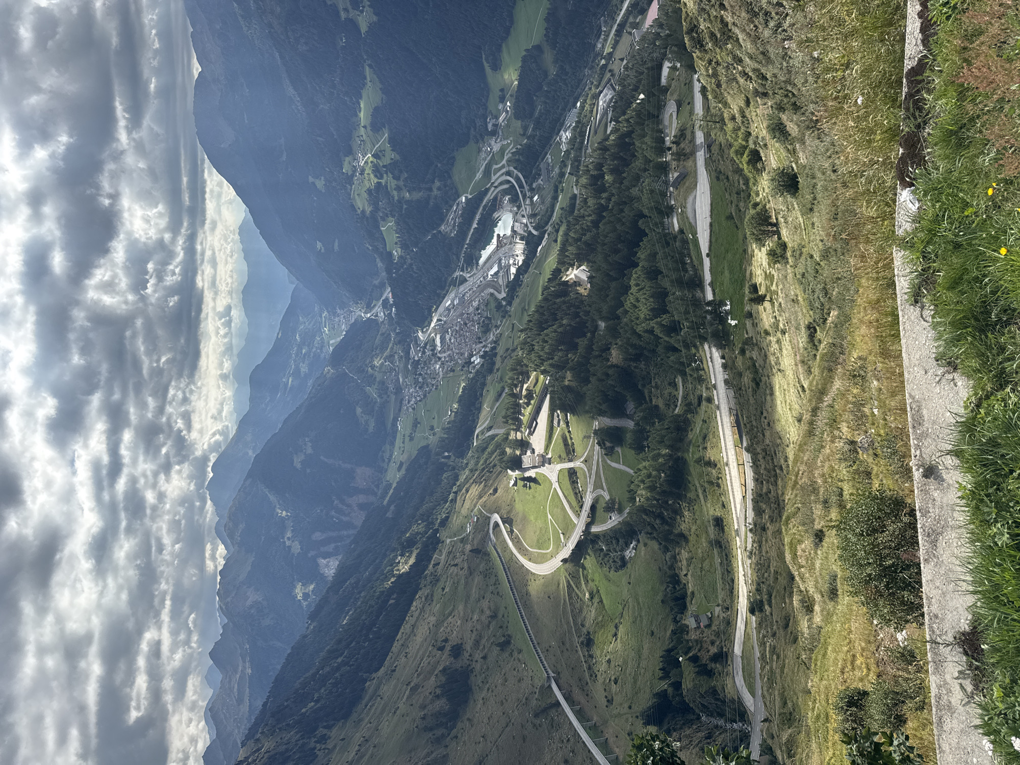The image depicts a panoramic view of the Swiss Alps, specifically capturing the St. Gotthard Pass area near Airolo, Switzerland. The scene is dominated by a dramatic mountainous landscape under a cloudy sky.
In the foreground, the terrain is grassy and uneven, with some rocks and sparse vegetation. A concrete barrier runs along the bottom of the image, suggesting a viewing platform or roadside.
The midground features a winding mountain road with multiple switchbacks, showcasing impressive engineering to navigate the steep slopes. The road appears paved and well-maintained, with some sections elevated on viaducts. Green meadows and scattered trees dot the landscape surrounding the road.
In the background, the valley opens up, revealing a town or village nestled amongst the mountains. This settlement appears to have a dense cluster of buildings, a lake or reservoir, and a complex network of roads and infrastructure, including what looks like a railway yard. Beyond the valley, numerous mountain ranges recede into the distance, their peaks softened by atmospheric perspective and the overcast sky.
The overall impression is one of grandeur and natural beauty, highlighting the challenging yet stunning terrain of the Swiss Alps and the human ingenuity involved in traversing it. There are no discernible people or specific actions taking place, focusing solely on the landscape and infrastructure.