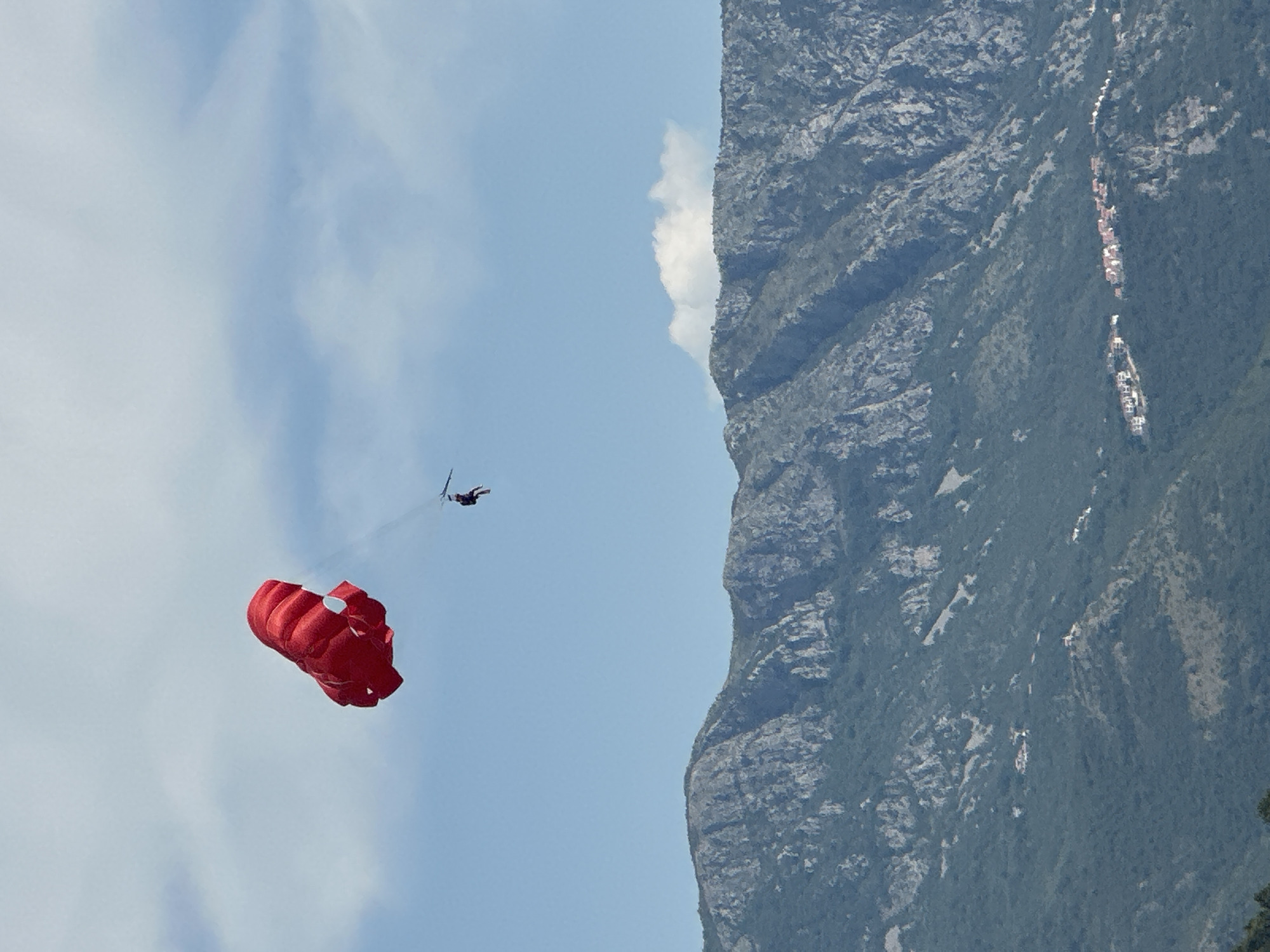 A red parachute with a person suspended beneath it is flying in a blue sky with scattered white clouds. The parachute is positioned in the upper left quadrant of the image. In the lower half of the image, a range of steep, rocky mountains is visible. Small clusters of white buildings with red roofs are scattered across the lower slopes of the mountains, indicating a settlement. The image appears to be taken from a distance, looking towards the mountains and sky. This view is consistent with the coastal areas surrounding Budva, Montenegro, where mountainous terrain meets the sea.