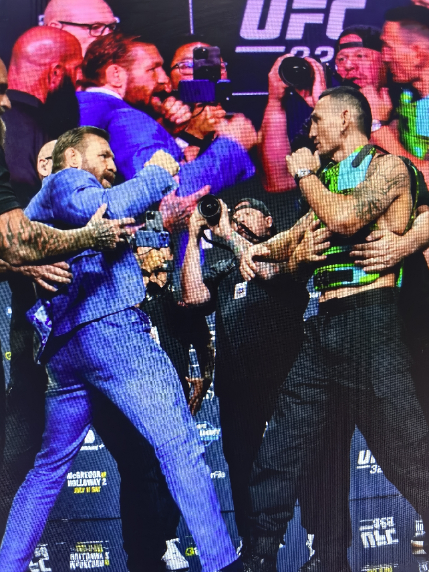 The image depicts a confrontational scene between two male athletes, likely fighters, at a UFC event. One man, wearing a blue suit, has his arms extended forward and appears to be lunging towards the other. The second man, wearing a green and black vest over black pants, has his arms raised and is holding a microphone. Both men have intense facial expressions. Surrounding them are several individuals, some holding cameras and phones, suggesting this is a press event or weigh-in. In the background, a large UFC logo is visible. The event appears to be taking place indoors on a stage, indicated by the lighting and the backdrop. There is no visual information in the image to suggest a location in Fergana City, Uzbekistan.