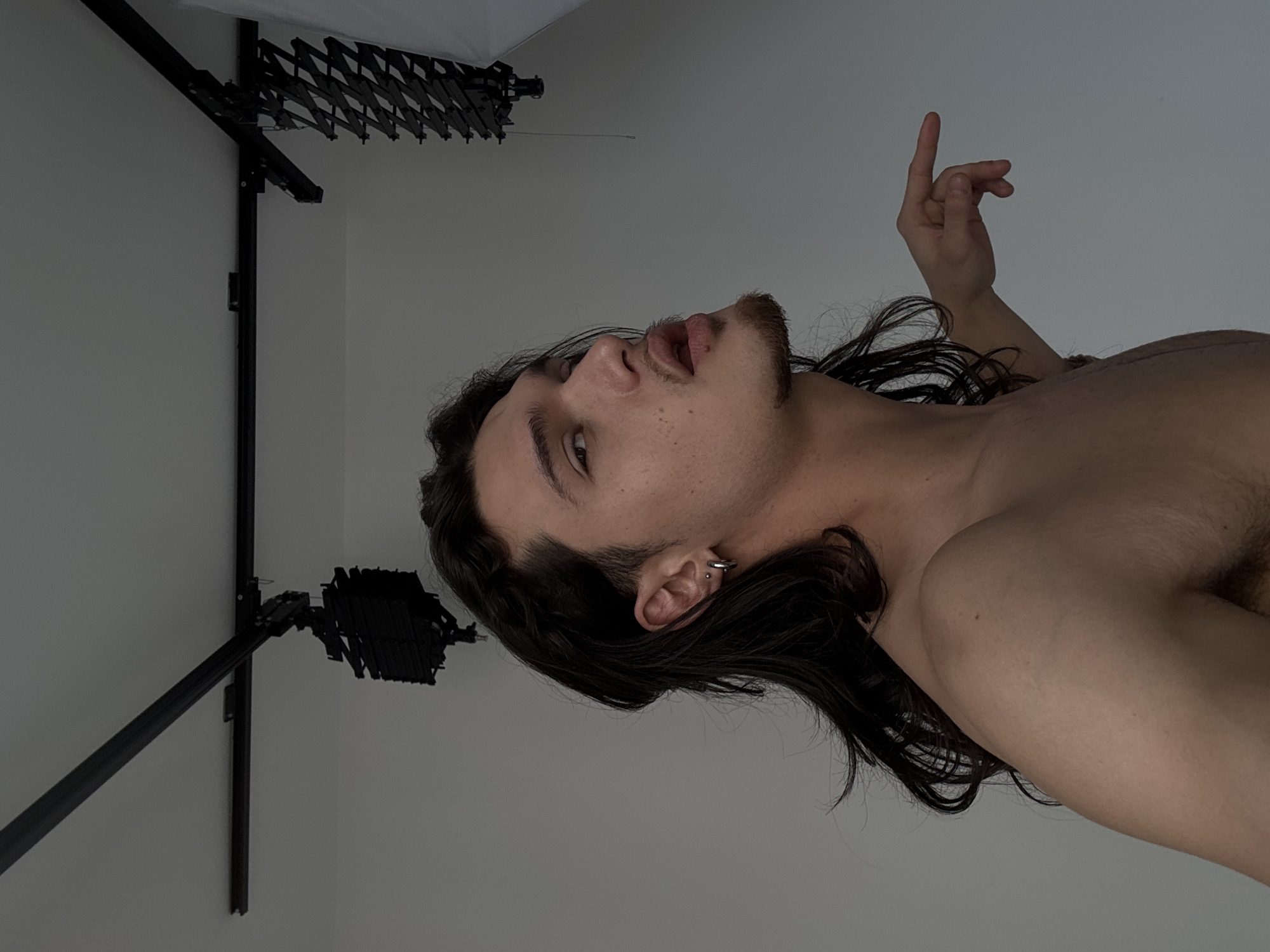 A young adult male, shirtless, is depicted from the upper torso upwards against a plain light gray background. He has long, dark, wavy hair, a goatee, and a thin mustache. His left ear features a silver hoop earring in the lobe and a small silver stud higher on the helix. His head is tilted back and slightly to the right, with his eyes looking towards the camera (viewer's left), and his mouth is slightly open. His right arm is raised, with the index finger extended and pointing horizontally to the right side of the frame. Above, a white ceiling features black metal tracks with professional studio lighting equipment, including a collapsed black pantograph arm and a partially visible white softbox on the right. The specific location within the city of Sofia, Bulgaria, cannot be determined from the provided image.