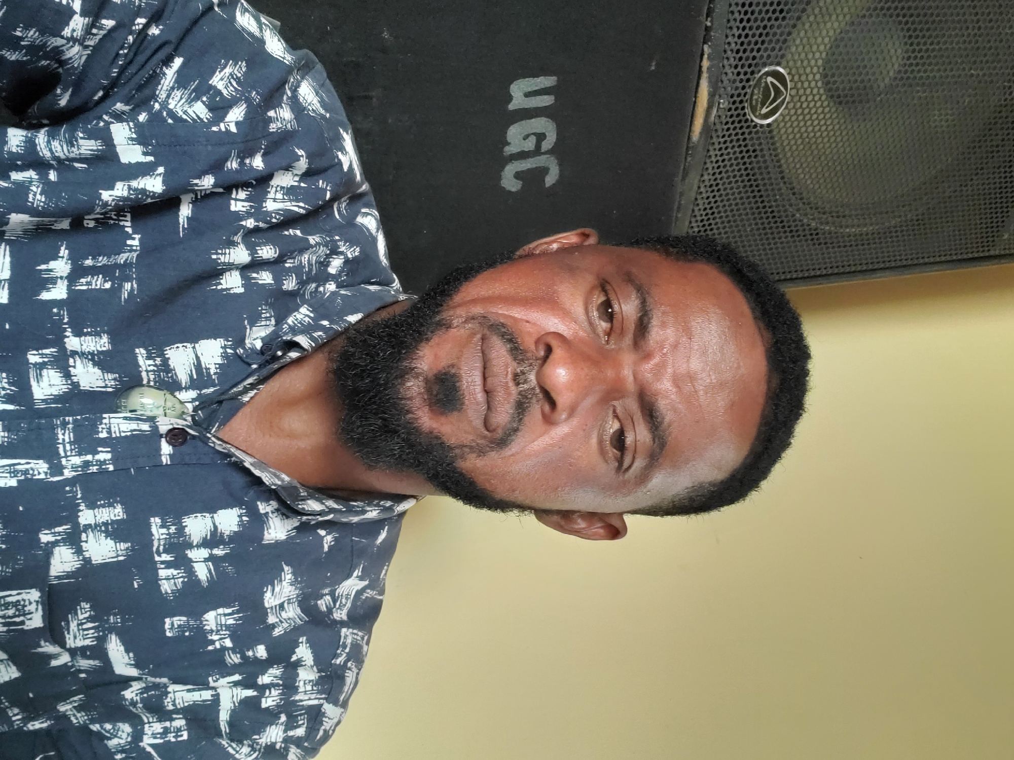 A medium shot captures a dark-skinned man with a black beard and mustache, wearing a dark blue and white patterned shirt. Behind him is a black speaker cabinet with the letters "ugc" printed on it, and to its right, another speaker with a metal grill. The background wall is a light yellow color. Given the provided context of "Jos, Nigeria," it is inferred the image was taken in this city.