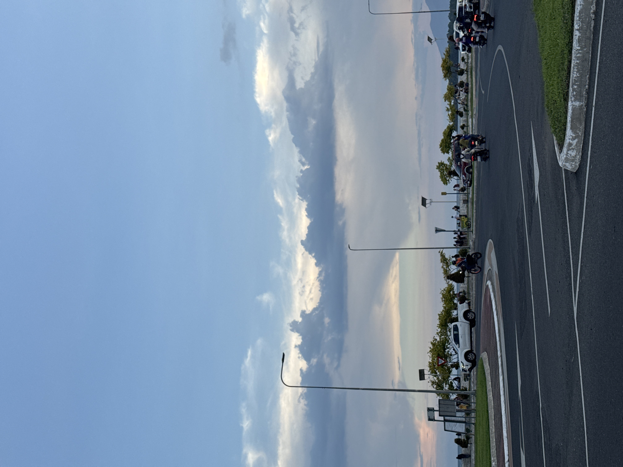 The image depicts a street scene in Davao City, Philippines. On the right, a curved road with white lane markings and a grass median is visible. A concrete barrier separates the median from the road. Motorbikes and a white pickup truck are parked along the road, oriented toward the left of the image. There are lampposts, trees, and several people standing near the parked vehicles. The sky is predominantly blue with some clouds.
