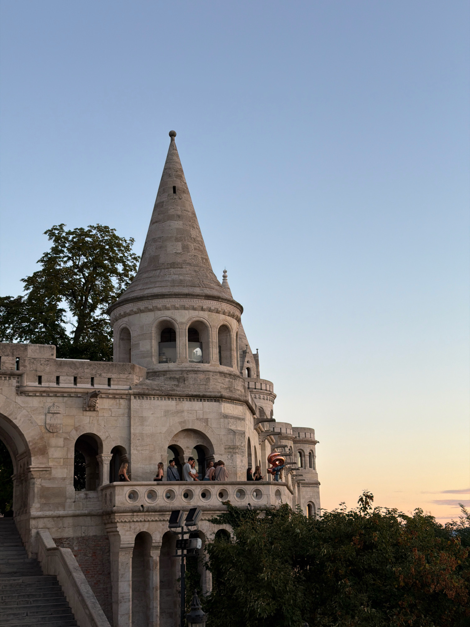The image shows a historic stone building with multiple turrets and balconies, characteristic of Fisherman's Bastion in Budapest, Hungary. The sky is clear and blue, transitioning to a pale orange hue at the horizon, indicating either sunrise or sunset. Several people are visible on a balcony, some standing and looking out, while others are engaged in conversation. One person appears to be holding a large, rose-gold colored balloon shaped like the number "8". Lush green trees are interspersed with the architecture, and a set of stone stairs is visible on the left side of the frame, leading up to the structure. The overall scene captures a peaceful moment with a blend of architectural grandeur and human activity against a natural backdrop.