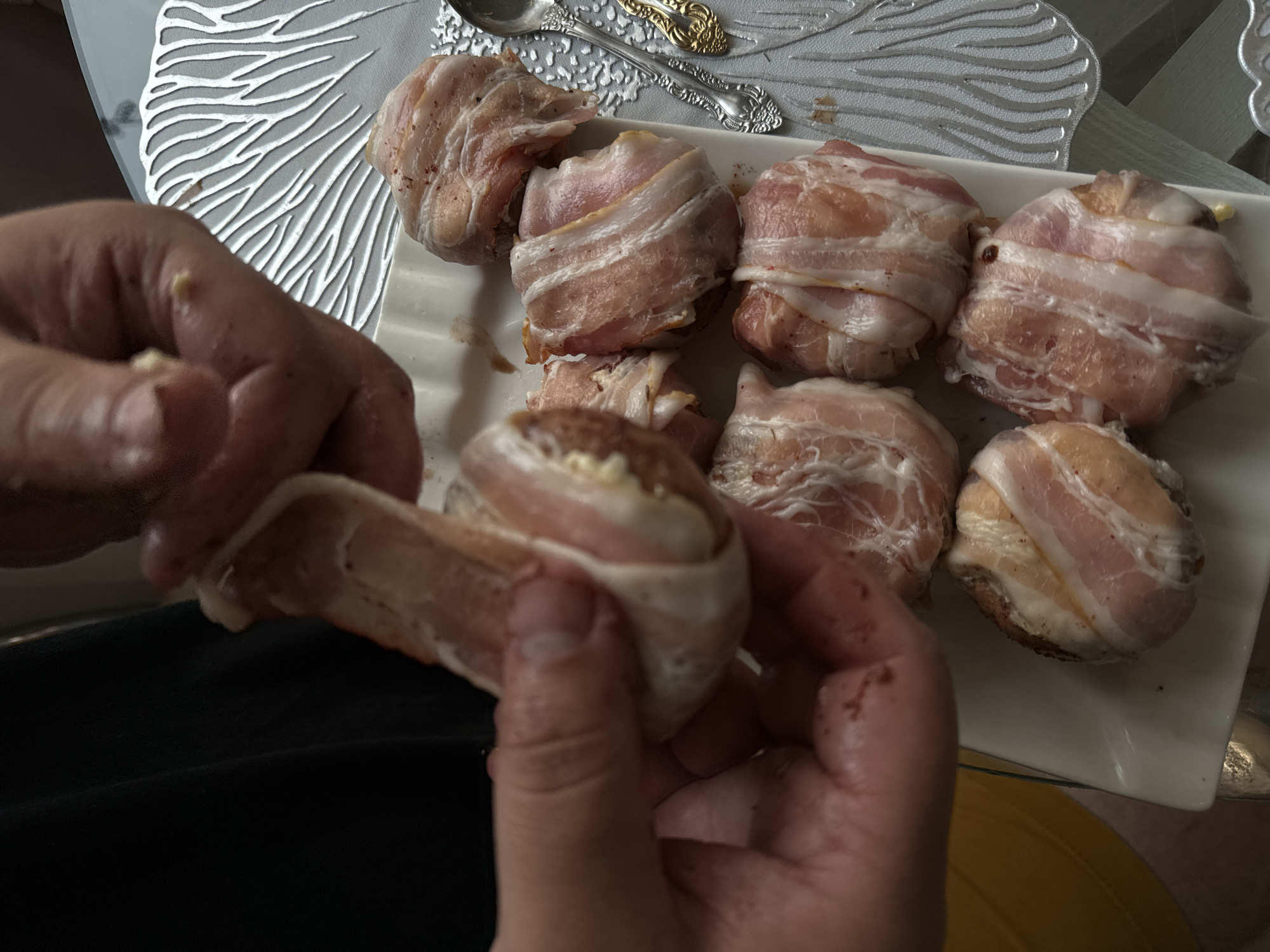 A person is preparing food, specifically wrapping small, round items in bacon. The items appear to be some type of meatball or appetizer. The person's hands are visible, one holding a piece of bacon and the other guiding it around one of the bacon-wrapped items. A white platter holds several other similarly prepared items. The scene is indoors, with a table and possibly a kitchen setting suggested by the background elements, including decorative placemats and cutlery. The lighting suggests it is daytime.