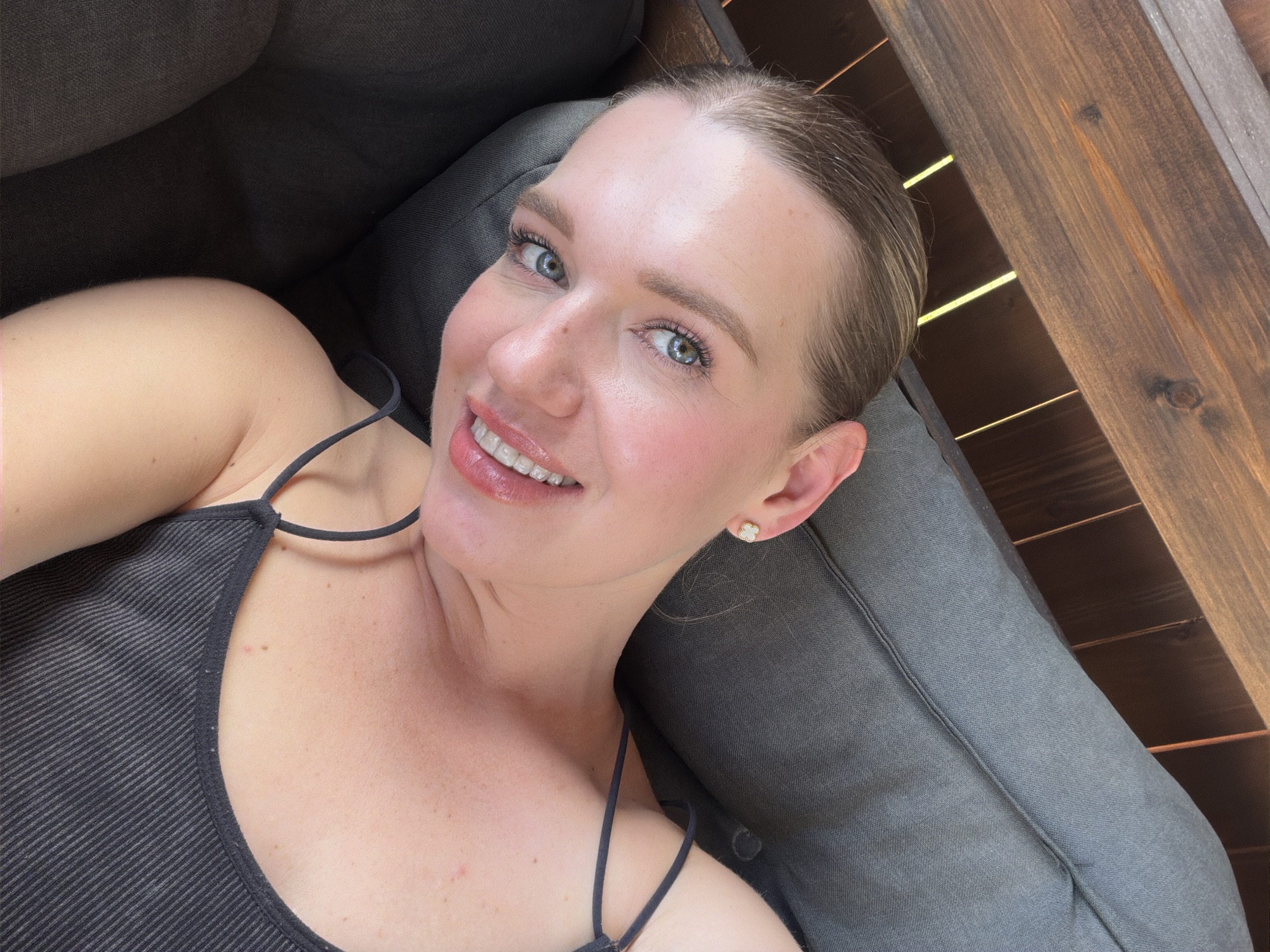 A female subject with light brown hair secured away from her face and blue-green eyes is depicted in a reclined position on dark grey upholstered outdoor furniture. She is smiling directly at the viewer, showing her upper teeth, and appears to be wearing light facial makeup. Her attire consists of a dark grey, ribbed tank top with thin spaghetti straps. A small, white, four-leaf clover-shaped stud earring with a gold center is visible in her right ear. In the background, behind her head, are stained dark brown wooden planks, consistent with outdoor fencing or a pergola, through which several vertical shafts of bright sunlight are visible. The scene is outdoors, located in Dreiliņi, Latvia.