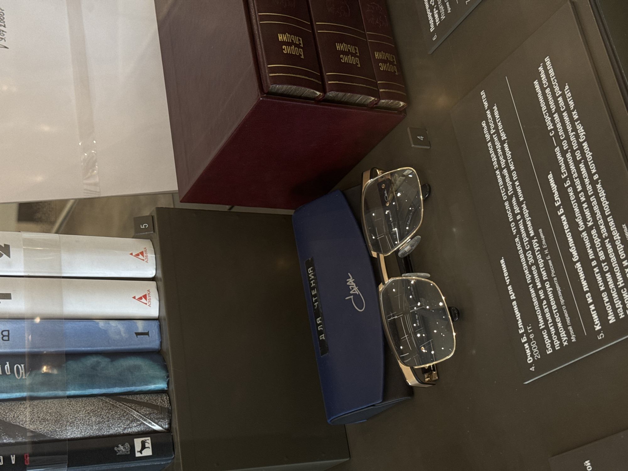 The image depicts a close-up view of a display case, likely within a museum setting. The focus is on a collection of books and a pair of glasses. The books are bound in a rich, dark red material with gold lettering on their spines, which appears to be in Russian. The visible text on the book spines reads "Борис Ельцин" (Boris Yeltsin).
Beside the books, a pair of modern eyeglasses with gold frames rests on a dark blue glasses case. The case has the word "CAZAL" printed on it in script, and the words "ДЛЯ ЧТЕНИЯ" (for reading) in block letters.
A printed informational placard is also visible, tilted diagonally across the frame. This placard provides text in Russian, detailing the objects. The text mentions "Очки Б. Ельцина для чтения" (B. Yeltsin's reading glasses) and discusses his reading habits after leaving office, stating he aimed to read at least 300 pages a day and enjoyed fiction, memoirs, history, and detective novels. It also refers to "Книги из личной библиотеки Б. Ельцина" (Books from B. Yeltsin's personal library) and mentions that many of these books have dedications from their authors. The placard indicates that the display is from the Museum of the First President of Russia, B. N. Yeltsin.
The overall scene suggests a curated exhibit honoring Boris Yeltsin, the first President of Russia. The lighting is diffused, indicating an indoor setting, possibly a museum. The arrangement of the items is neat and organized, as is typical for displays of historical artifacts. There are no indications of weather or time of day, but the environment appears well-lit and controlled.