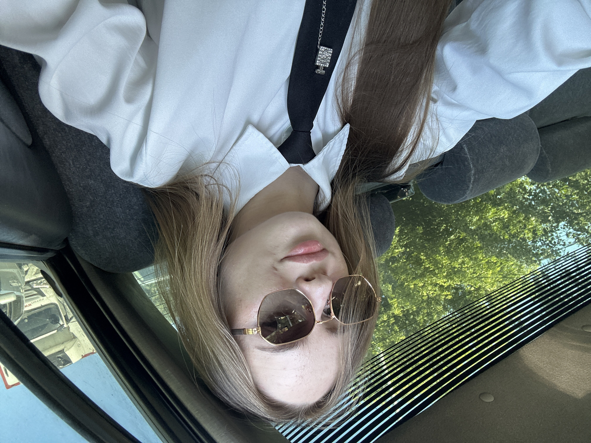 A young woman with long blonde hair and fair skin is taking a selfie inside a car. She is wearing a white button-down shirt, a black tie with a jeweled perfume bottle clip, and oversized sunglasses. The car's interior is visible, including dark grey seats and the rear window with horizontal blinds. Outside the window, green trees and a bright blue sky can be seen. In the background, through the car's side window, a white truck is visible. The lighting suggests it is daytime.