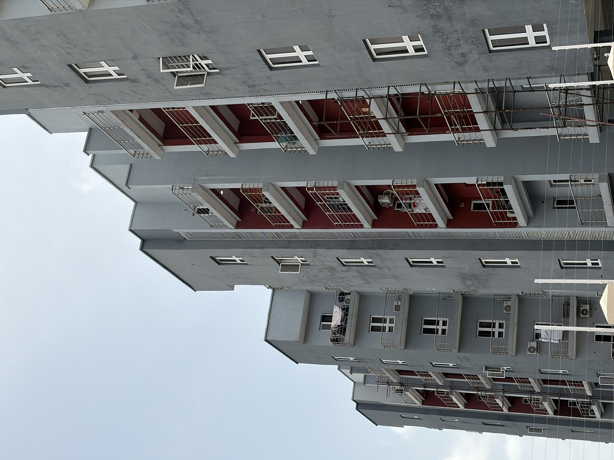 The image shows an apartment building in Igboefon, Nigeria. The building has multiple floors, with a gray exterior and red balconies. Windows are rectangular. Some balconies have metal railings. Air conditioning units are mounted on some exterior walls. There are clothes hanging from a balcony. The sky is visible in the background. Power lines are strung between buildings.
