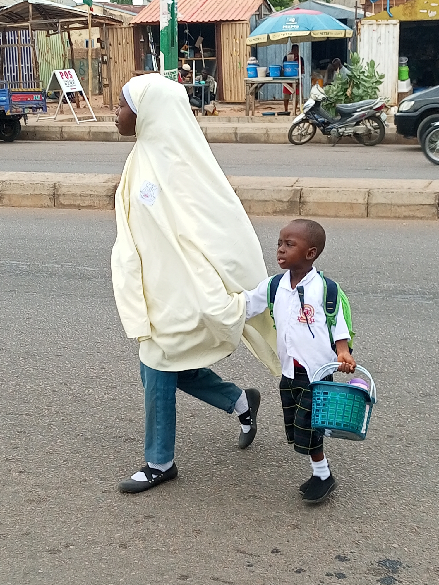 A medium, eye-level shot depicts a woman and a young boy walking on a paved street in Tempure, Nigeria. The woman, wearing a long, cream-colored hijab, blue jeans, and black shoes with a strap, is on the left side of the frame. The boy, dressed in a white shirt, plaid shorts, white socks, and black shoes, is carrying a green and blue plastic basket and has a green backpack on. They appear to be crossing the street.
In the background, there are various structures including a small business with a "POS" sign, a motorcycle parked near a blue umbrella-covered food stand, and a dark car. Other people are visible in the background, near the structures. The sky is overcast.