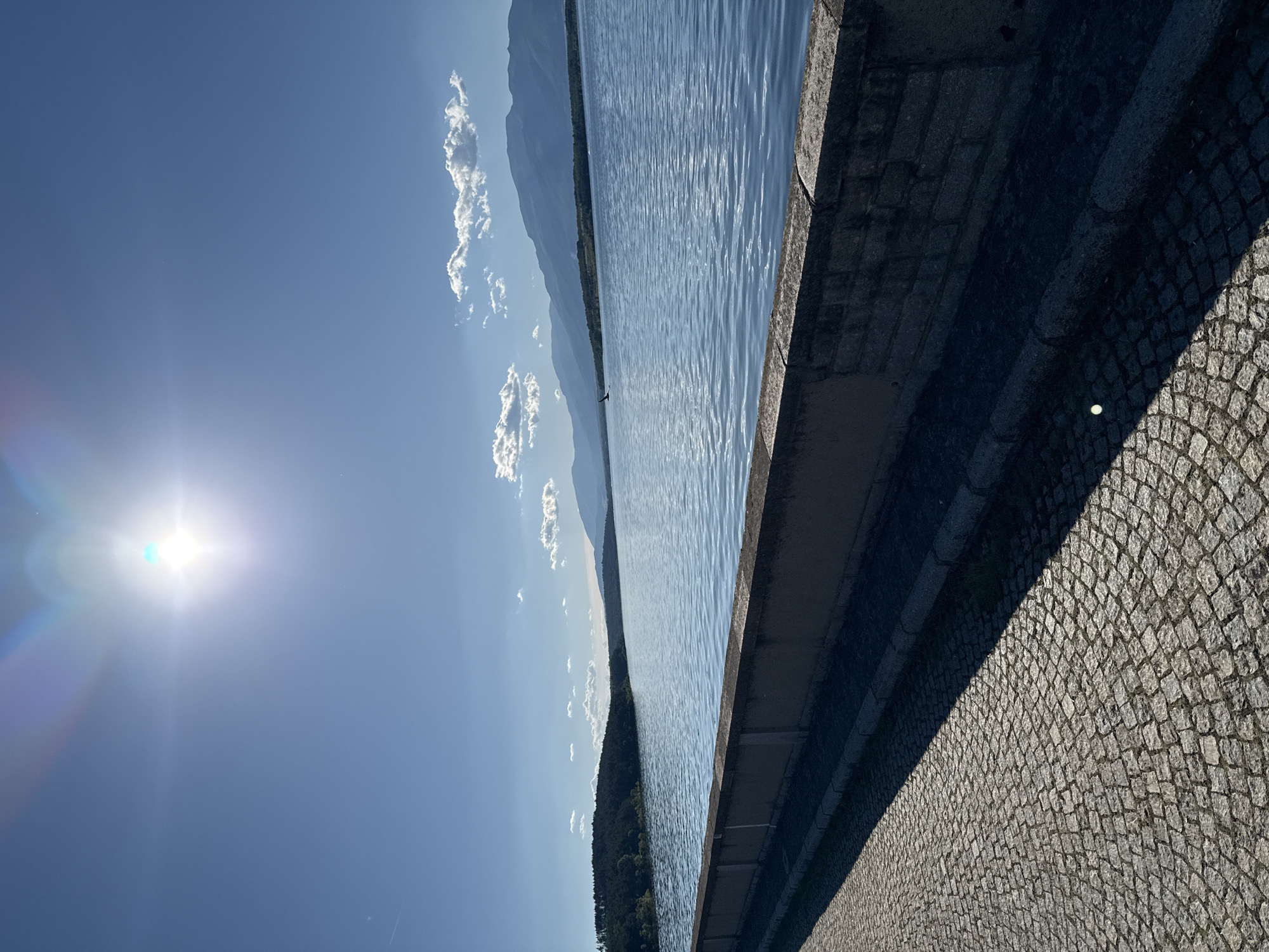 A sunlit, wide-angle shot shows the Koprinka Dam in Koprinka, Bulgaria. The foreground features a cobblestone pathway with a distinct shadow cast by a wall that runs along the edge of the dam. The wall is constructed from stone blocks and has a concrete cap. Beyond the wall, the vast, calm surface of the reservoir reflects the bright blue sky and the sun, which is high and slightly off-center, creating lens flare. The water shows gentle ripples. In the middle ground, a tree-covered hill slopes down to the water's edge on the left. Further in the distance, a range of hazy mountains forms the horizon line. A single bird is captured in flight over the water. The sky is clear with a few scattered, fluffy white clouds.