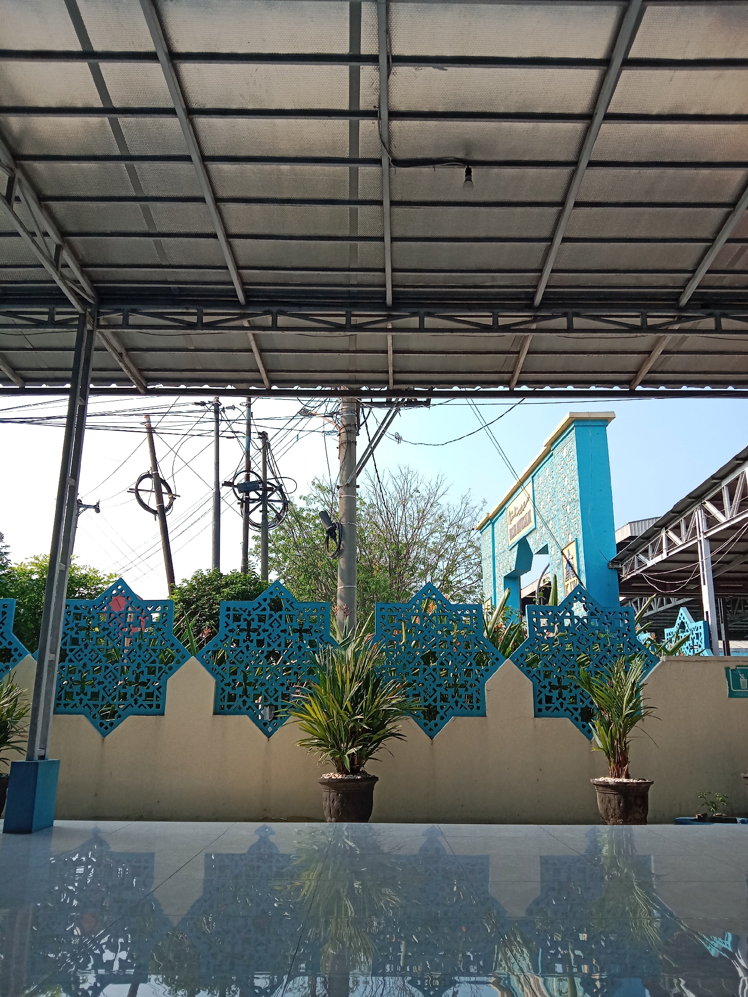 The image captures a view from under a metal roof structure, looking out towards a building and its surroundings. In the foreground, a polished floor reflects the scene above, creating a mirror image of the overhead structure and the environment beyond. A cream-colored wall with a decorative blue lattice runs horizontally across the midground, behind which are potted plants with lush green foliage. To the right of the wall, a bright blue building with a sign is visible, suggesting it might be a mosque or a public facility. The blue building has an arched entrance and is adorned with what appears to be a stone-like texture. Further to the right, another covered structure with a metal roof is partially visible. Power lines and utility poles are prominent in the background, indicating an urban or semi-urban setting. The sky is clear and blue. This scene is located in the city of Semarang, Indonesia, likely at the entrance of a mosque.