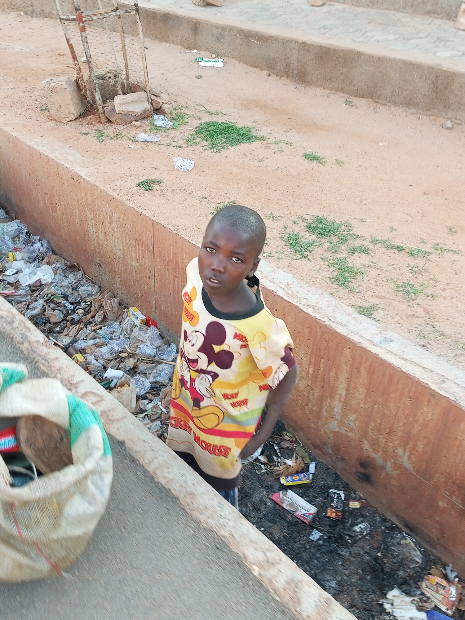 In Tempure, Nigeria, a young boy stands in a garbage-filled ditch. He is wearing a yellow t-shirt with Mickey Mouse and other cartoon characters printed on it. The ditch is lined with concrete and filled with plastic bags, paper, and other debris. A woven bag filled with various items, including what appears to be a coconut, is visible to the left of the boy. The surrounding area is sandy with patches of dry grass. In the background, there is a rusty metal fence and concrete blocks.