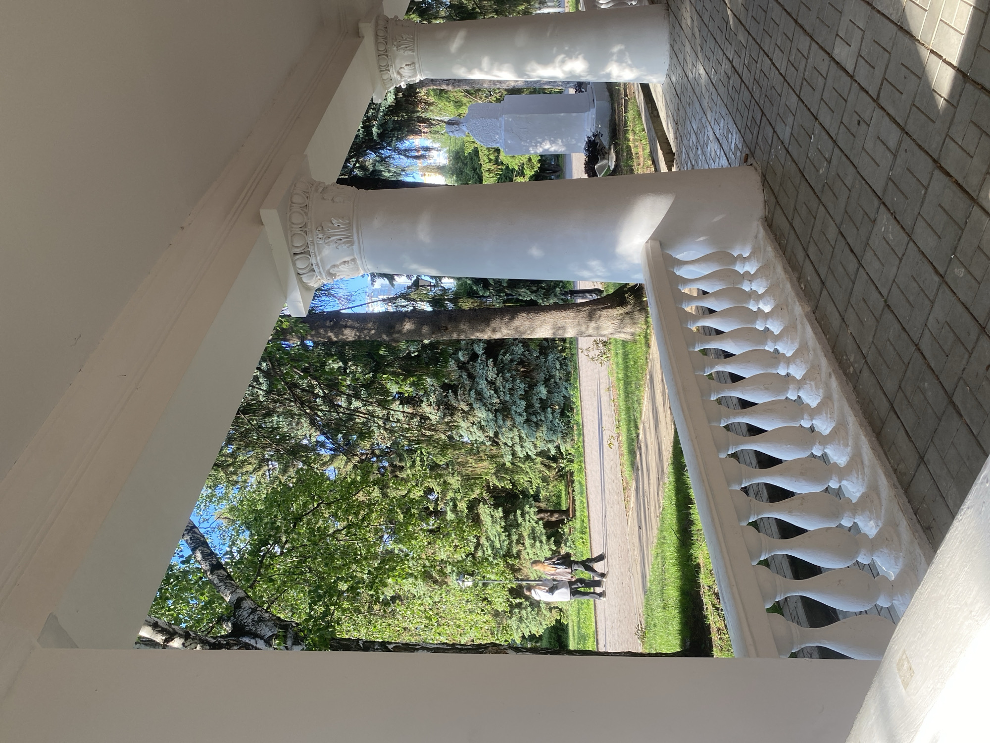 The photograph depicts a view from a white, classical-style portico. Fluted columns with ornate capitals support the structure. A white balustrade runs along the edge. Outside the portico, a green lawn extends towards a paved walkway. Two people are walking on the path. Trees and foliage are visible. Further in the background, a white structure is present. The location is likely the Kharitonov-Rastorguyev Estate in Yekaterinburg, Russia, due to its distinctive architectural features. The scene suggests a sunny day.