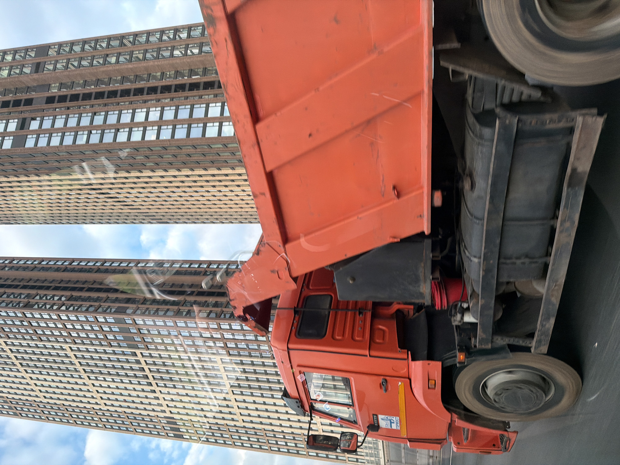 This media file, originally rotated 90 degrees counter-clockwise, depicts an urban scene with an orange dump truck in motion alongside two tall skyscrapers, under a partly cloudy sky.
The primary subject is a large, bright orange dump truck, occupying the right and lower portions of the frame (when viewed upright). Its wheels are blurred, indicating movement. The truck's cabin is visible, featuring a large side mirror and several small stickers. On the side of the cabin, the number "55111" is clearly visible, along with the text "УРАЛМАШ" (URALMASH) and "ТОН" (TON), likely referring to the manufacturer and capacity. The dump bed, also orange, shows signs of wear and scratches. The undercarriage of the truck, including a fuel tank and mechanical components, is also visible.
In the background, two prominent skyscrapers rise against a blue sky with white clouds. These buildings are modern, featuring a distinctive brown or beige facade with numerous rectangular windows that reflect the sky. The closest building appears to have a textured or grid-like pattern across its surface. The buildings suggest a contemporary city environment.
The photo appears to be taken during the daytime, likely on a sunny or partly cloudy day, given the lighting and visible sky. The setting is an urban landscape, consistent with the provided location context of Moscow, Russia. The motion blur on the truck's wheels suggests the photograph was captured from another moving vehicle or rapidly from a stationary point as the truck passed. No people are explicitly visible in the image, and no specific emotions are conveyed.