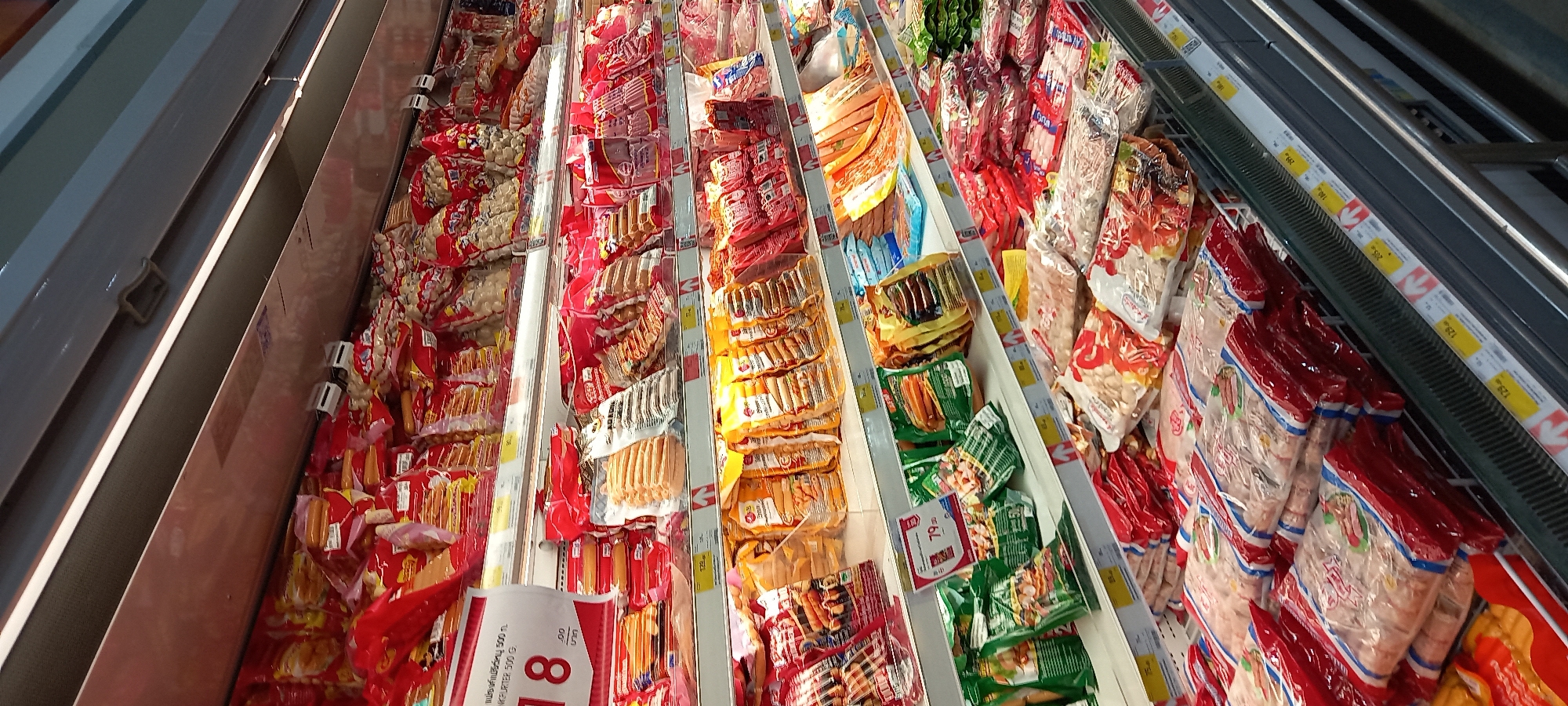 This media file depicts an overhead view of a supermarket or grocery store freezer aisle in Moo 1, Thailand. The primary subjects are numerous packaged food items, predominantly different varieties of sausages, hot dogs, and other processed meat or fish products. These items are displayed within long, multi-tiered refrigerated or freezer cases.
The activity taking place is the commercial display of food products for sale. The items are organized and stacked in individual clear plastic packages, many featuring vibrant red, green, or yellow branding and labels. The display cases are well-lit, highlighting the products.
Notable details include the extensive variety of similar products, suggesting a specialized section for these items. Several price tags are visible along the edges of the display cases. Visible text includes prices such as "129", "299", "189", "205", and "79.00". A larger red and white sign prominently features a large "8" and "79.00". One product label clearly shows the Thai text "แฟรงค์แม่ชีร์หนู 500 ก." (Frankfurter 500 g.) and the English text "NKFURTER 500 G.". No people are visible in the image, and the time of day is not discernible.