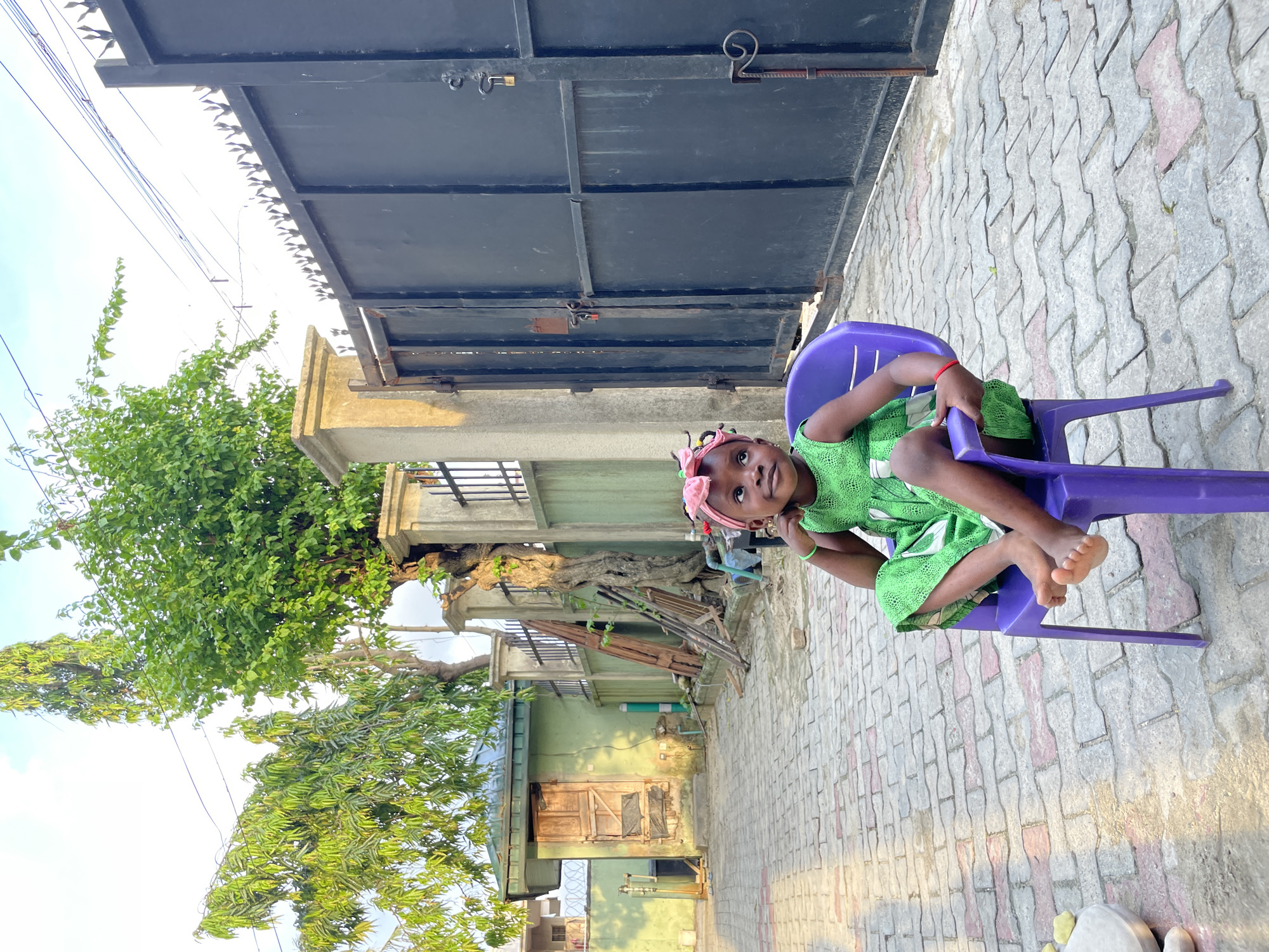 This image captures an outdoor scene, specifically described as being in the city of Ado, Nigeria. The foreground consists of an interlocking paver surface, primarily composed of grey blocks with interspersed reddish-brown elements.
In the midground, a young girl is seated facing slightly left on a small, purple plastic chair. She wears a vibrant green sleeveless dress and a pink fabric headband adorned with small, colorful beads or decorations in her hair. Her bare feet are resting on the seat of the chair, with her legs bent. A red bracelet is visible on her left wrist and a green bracelet on her right wrist. Her right arm is bent with her hand near her chin, and she is gazing upwards and slightly to her left with an attentive expression.
The background features various architectural and natural elements. To the left, a small, light green utility or ancillary building with a dark, possibly corrugated metal, roof is visible. It has a rustic, weathered wooden door with visible horizontal slats and patches, and a small, boarded-up opening to its left. Adjacent to this building, a large tree with a thick, gnarled trunk and dense green foliage rises behind a light-colored concrete pillar. Wooden planks or a ladder are leaning against a low wall or fence near the tree.
To the right, a substantial dark-colored metal double gate, topped with decorative pointed spikes, is a prominent feature. Multiple padlocks are visible securing the gate panels. Adjacent to the gate are additional concrete pillars and sections of a light green wall or fence. Overhead, several utility wires or cables traverse the clear, bright sky. Further trees with various shades of green foliage are visible in the upper background, extending above the structures.