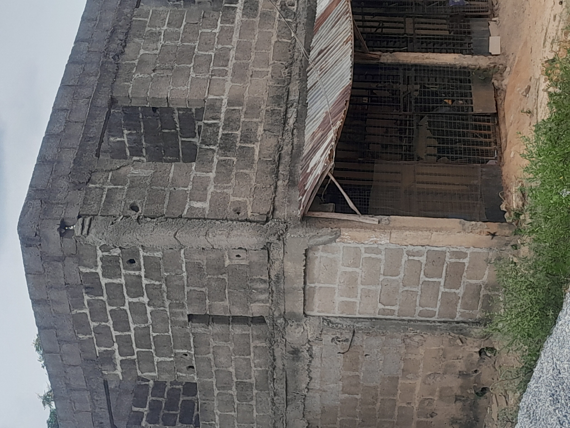 The scene depicts a two-story concrete block structure in Ifo, Nigeria, under an overcast sky. The building is constructed from unrendered grey concrete blocks, exhibiting an unfinished state or significant disrepair. The upper floor features multiple rectangular openings consistent with unglazed windows. A prominent, roughly finished horizontal concrete beam separates the ground and upper levels. The lower left side of the building contains a blocked-up opening and a doorway filled with concrete blocks. Extending from the lower right side is a rusted, corrugated metal awning, supported by unpainted wooden posts. Beneath this awning, multiple dark metal security grates or gates are visible, suggesting closed entrances or storefronts. The ground directly in front of the structure is unpaved earth, covered with patches of green weeds and scattered debris. A pile of light-colored gravel occupies the lower-left foreground. No individuals are present or engaged in any discernible activity within the frame.