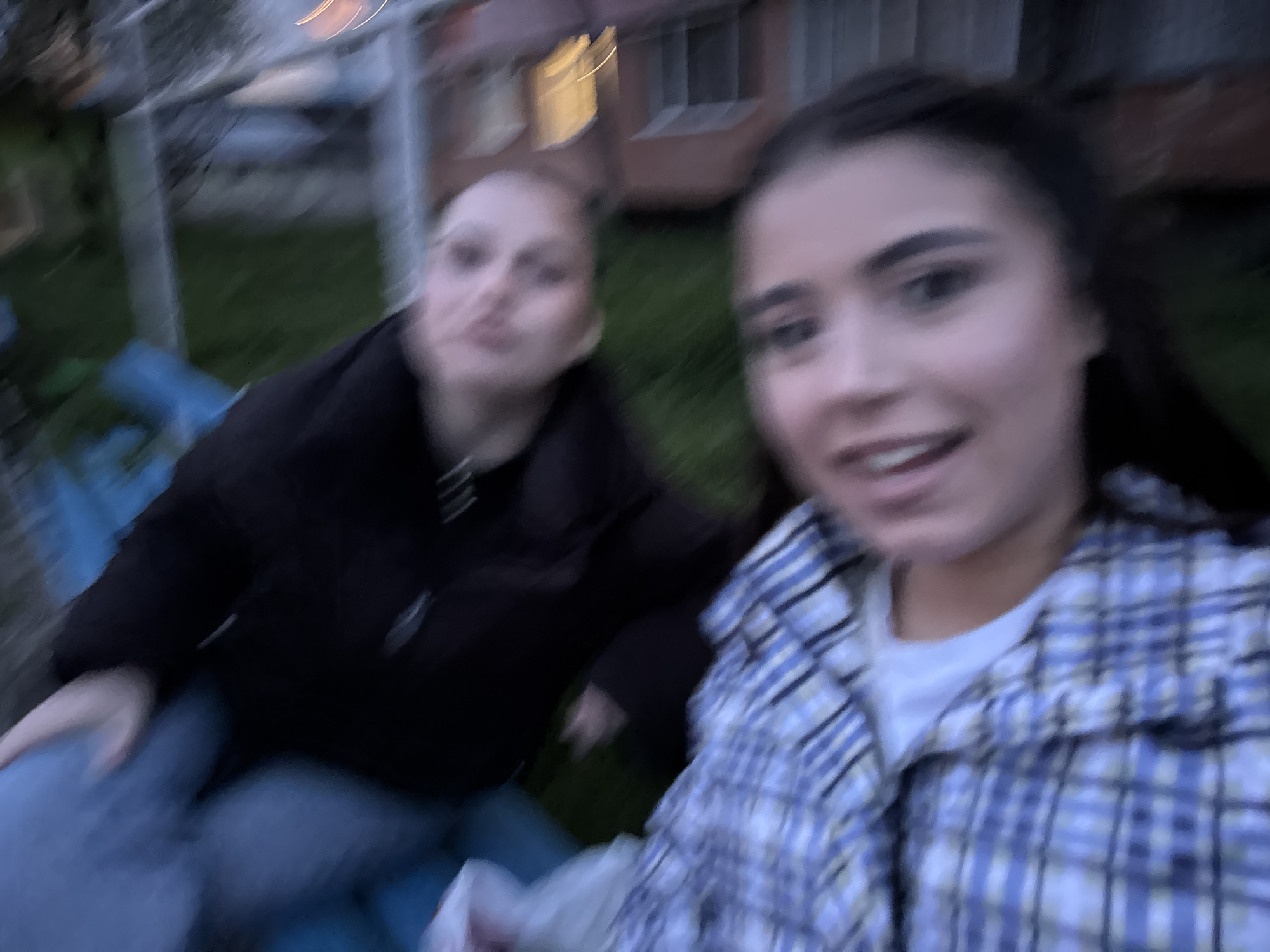 Two females are in the image, one is in the foreground on the right, wearing a light-colored plaid shirt over a white t-shirt, and the other is in the background on the left, wearing a dark jacket. They appear to be outdoors; in the background, there is green vegetation and the corner of a building. It's difficult to determine the exact location in Sofia, Bulgaria without more context. The quality of the image is blurry, giving the impression that the photo was taken in motion.