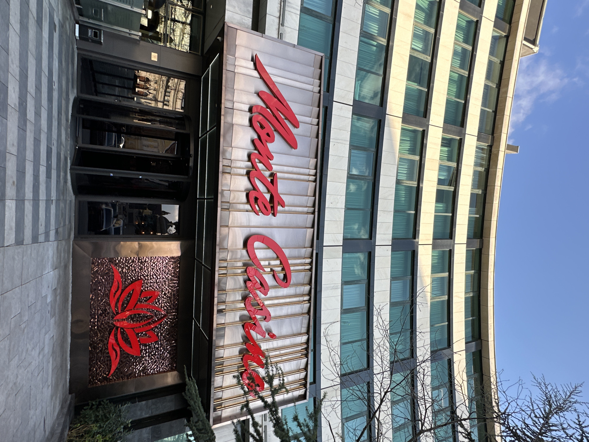 The image shows an exterior view of the Monte Casino in Sofia, Bulgaria. The casino's sign is prominently displayed in red lettering against a multi-tiered, metallic background. The sign reads "Monte Casino." To the left, there's a decorative, red floral design against a textured surface. The building's facade features a modern design with multiple windows. A clear blue sky is visible in the background. Bare branches of trees can be seen in the lower right corner of the image. The entrance area with glass doors and reflective surfaces is also visible.