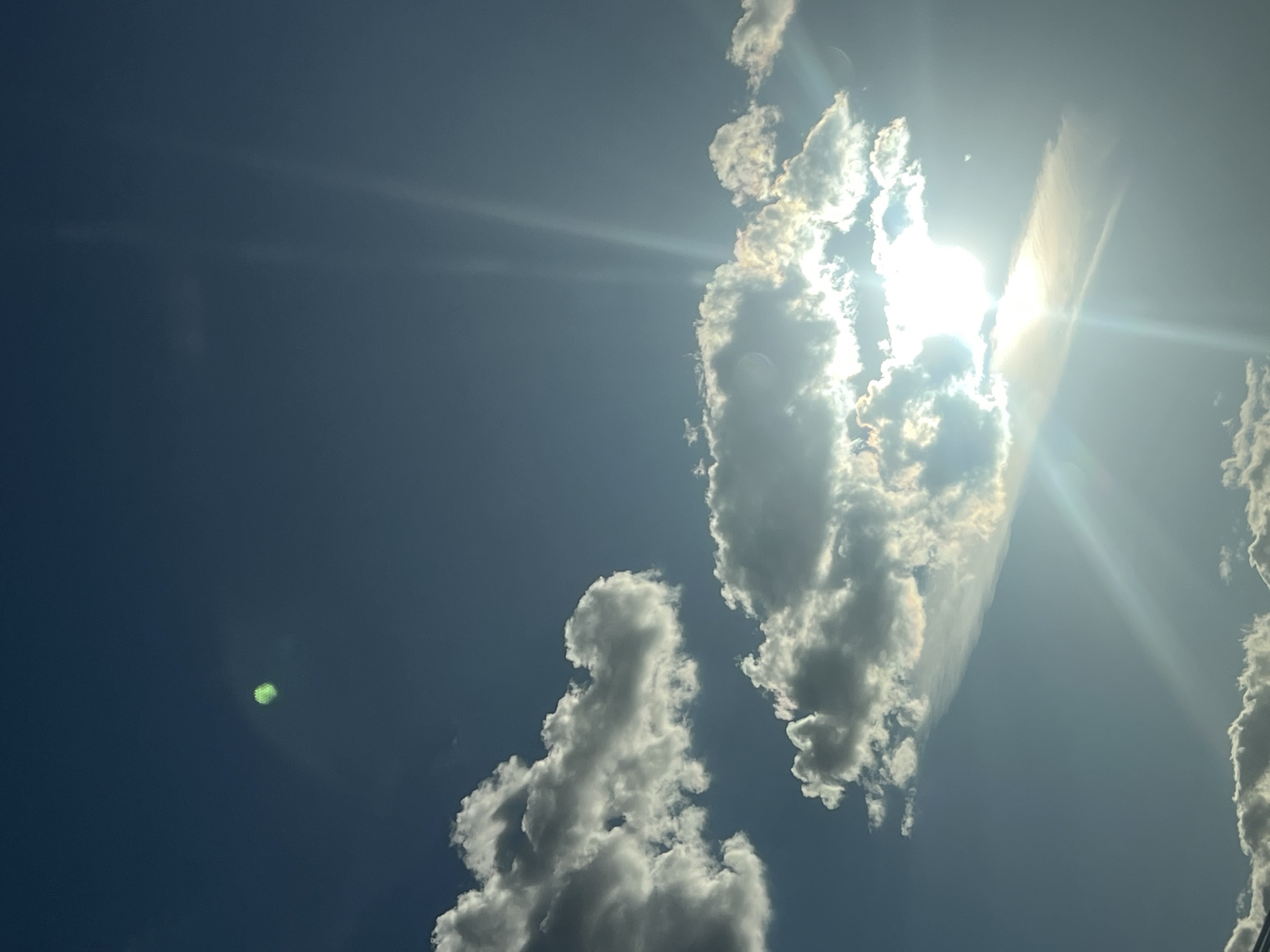 The media file shows a sky with clouds and the sun. The sun is bright and flares are visible, indicating a clear day. There are several cumulus clouds scattered across the sky. In the lower left corner, there is a distinct green, hexagonal lens flare. In the upper right, a long, thin cloud stretches across the frame. The overall mood is bright and airy, typical of a sunny day. No text is visible in the image. The setting is an outdoor sky scene, likely Moscow, Russia, given the provided location context. The time of day is clearly daytime, with the sun high in the sky.