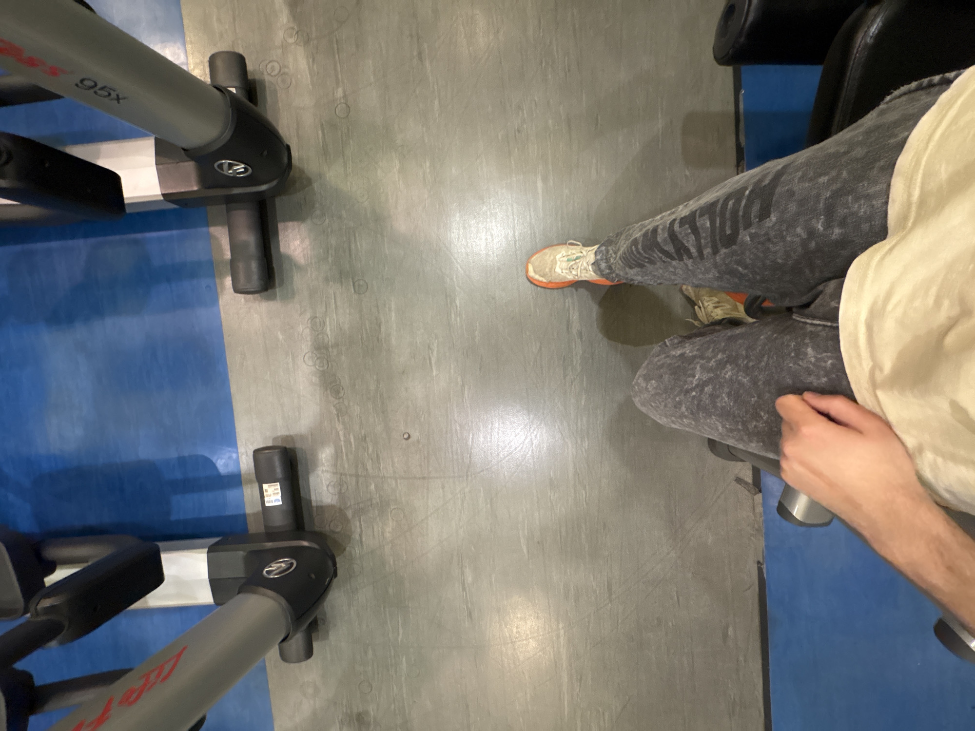 The image shows an overhead view of a person sitting on an exercise machine, possibly in a gym, although the location is not definitively identifiable as Mexico City. The person is wearing acid-wash grey jeans, a tan shirt, and orange-soled sneakers. The individual is holding onto the armrest of the machine. Two pieces of exercise equipment, labelled "95x" and "Life Fitness," are visible in the image. The floor is grey with a blue area surrounding the exercise machines.
