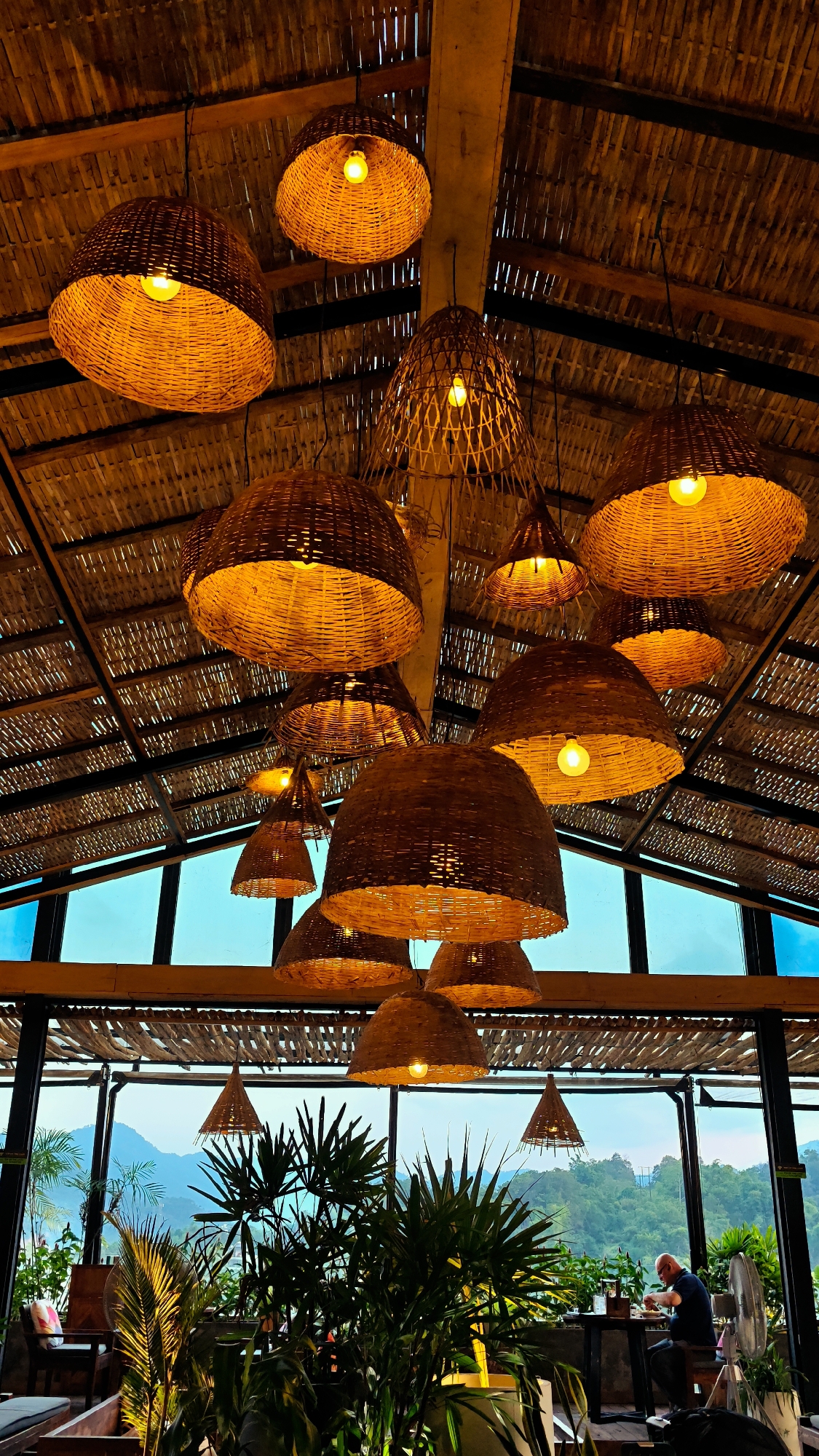 The image depicts the interior of a restaurant or cafe in Hà Giang 1 Ward, Vietnam. The most prominent feature is the ceiling, constructed from bamboo and adorned with numerous woven wicker light fixtures. Natural light filters through large windows, offering a view of a landscape with plants and mountains. A seated man eats alone at a table with a fan nearby. Other seating areas are visible with cushions, and plants decorate the foreground, adding greenery to the indoor space.