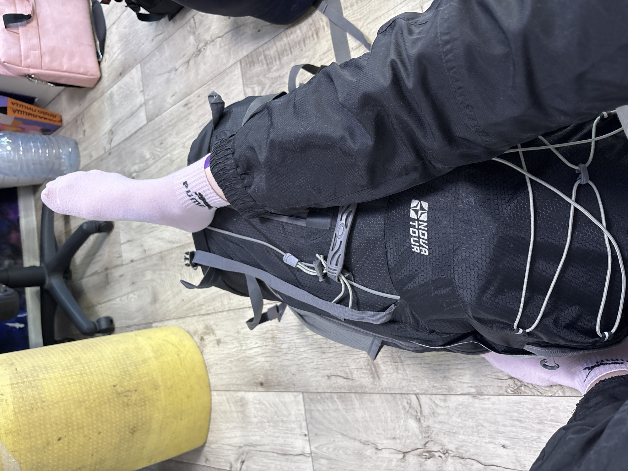 The image, taken in Прикамский, Russia, presents a high-angle, close-up view of a person's lower legs and feet in an indoor setting. The individual is wearing black pants and light pink socks, one of which clearly displays the "Puma" logo on the ankle. One leg is propped up on a large black backpack. This backpack has grey straps, white bungee cords on its front, and a textured hexagonal pattern. The brand "ENDUR TOUR" is visible on the upper right side of the backpack.
The setting features light-colored wooden or laminate flooring. In the background, a pink rectangular bag, possibly a laptop bag, is situated next to a clear plastic water bottle. To the far left, part of a black office chair's base and wheels can be seen. A yellow cylindrical object, which might be a rolled-up mat or cushion, occupies the lower-left foreground. The scene suggests a moment of rest or preparation, possibly for travel, given the presence of the backpack and other bags. No specific time of day or emotions are discernible.