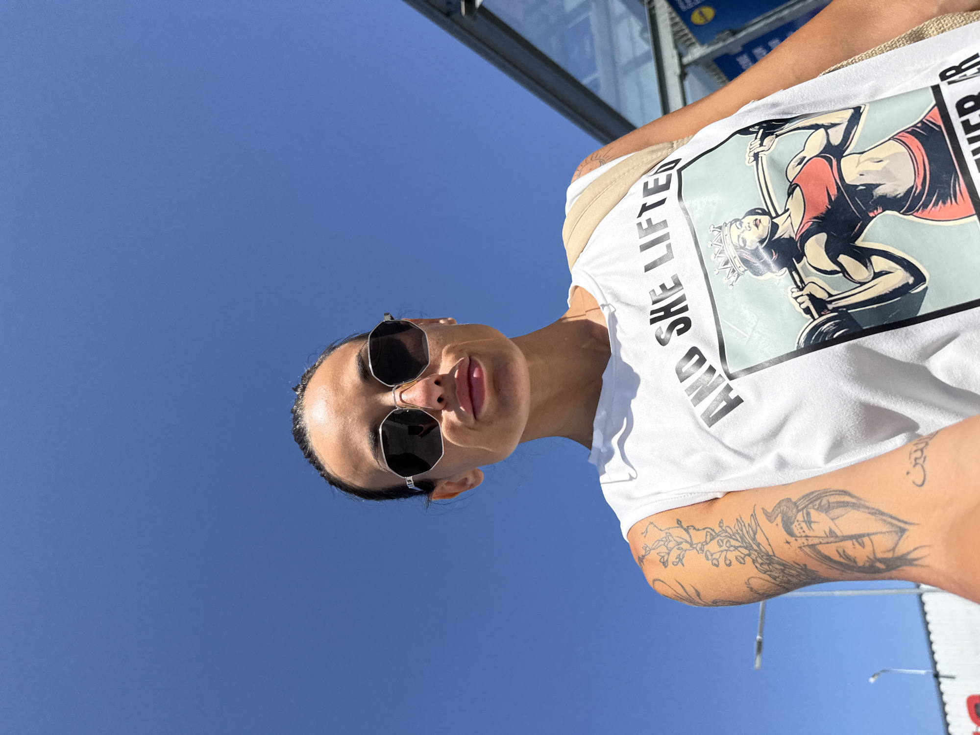 A woman with dark hair pulled back and wearing sunglasses takes a selfie against a clear blue sky. She is wearing a white tank top with a graphic of a muscular woman lifting weights and the text "AND SHE LIFTS" and "NEVER AR." The woman has tattoos on her left arm, including an image of two faces and a floral design. The background includes a glass structure and a street lamp, suggesting an urban environment. This image was taken in Pleven, Bulgaria.
