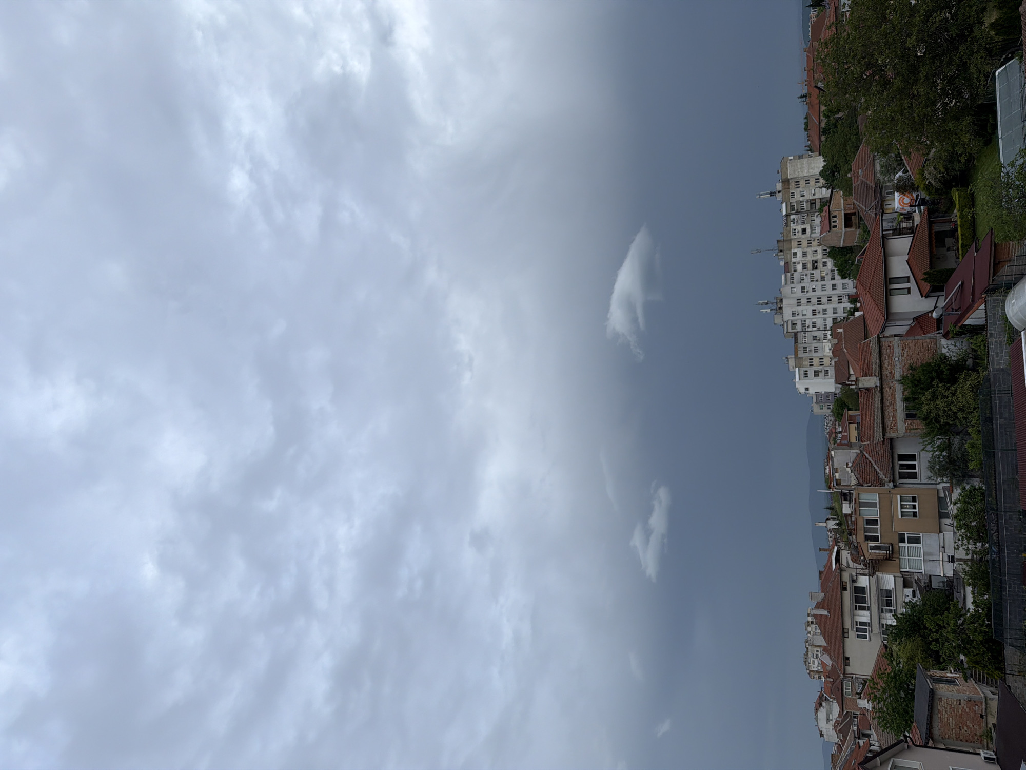The image presents a view of Burgas, Bulgaria, featuring a predominantly cloudy sky that occupies a large portion of the frame. The city skyline is visible on the right, revealing a mix of residential buildings with red-tiled roofs and taller apartment blocks. Trees are interspersed among the buildings. The overall atmosphere appears overcast, suggesting possible inclement weather.