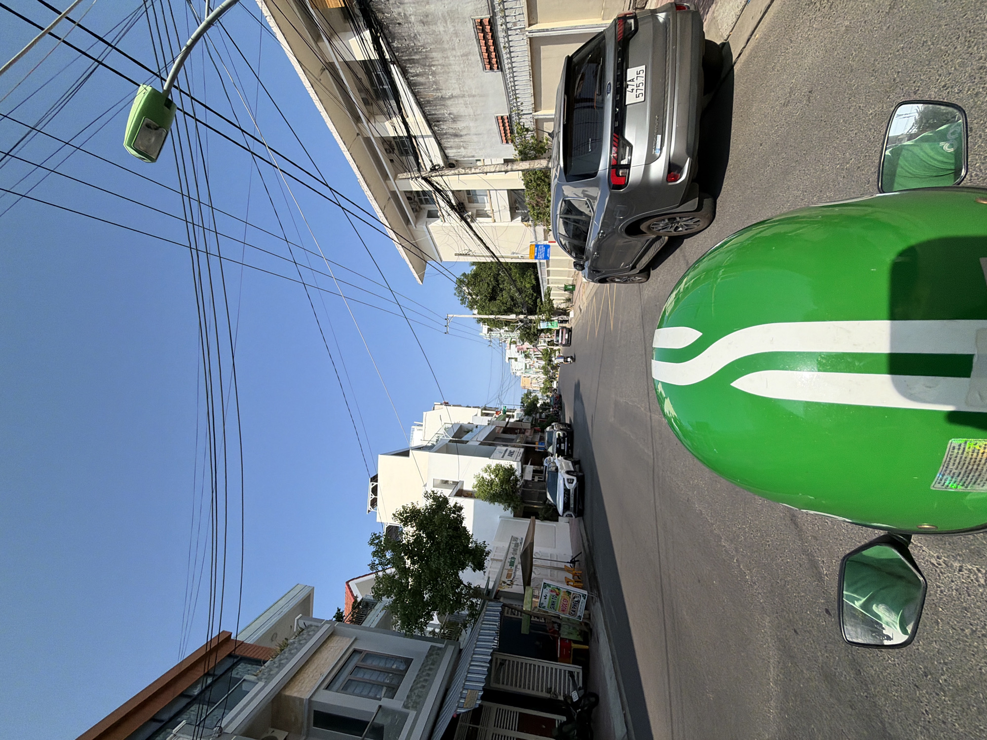 The image captures a street scene in Nha Trang, Vietnam, likely taken from the perspective of a passenger on a motorcycle. The foreground is dominated by the bright green helmet of the rider, with a white stripe running down the center. A side mirror reflects a person wearing a green shirt, presumably the rider. The street itself is paved and appears to be a typical urban road with buildings lining both sides.
On the left side of the street, several buildings are visible, including a multi-story structure with balconies and awnings. A parked motorcycle is also present near the curb. Further down the street, a white SUV and a dark-colored car are seen.
On the right side of the street, a large, light-colored building with multiple windows and air conditioning units is prominent. A concrete wall with a decorative fence runs along the property line. Further down the street, more buildings and a street lamp with a green fixture are visible. The sky is clear and blue, indicating a sunny day. The overall impression is of a bustling city street in Vietnam, with a blend of modern architecture and everyday urban life.