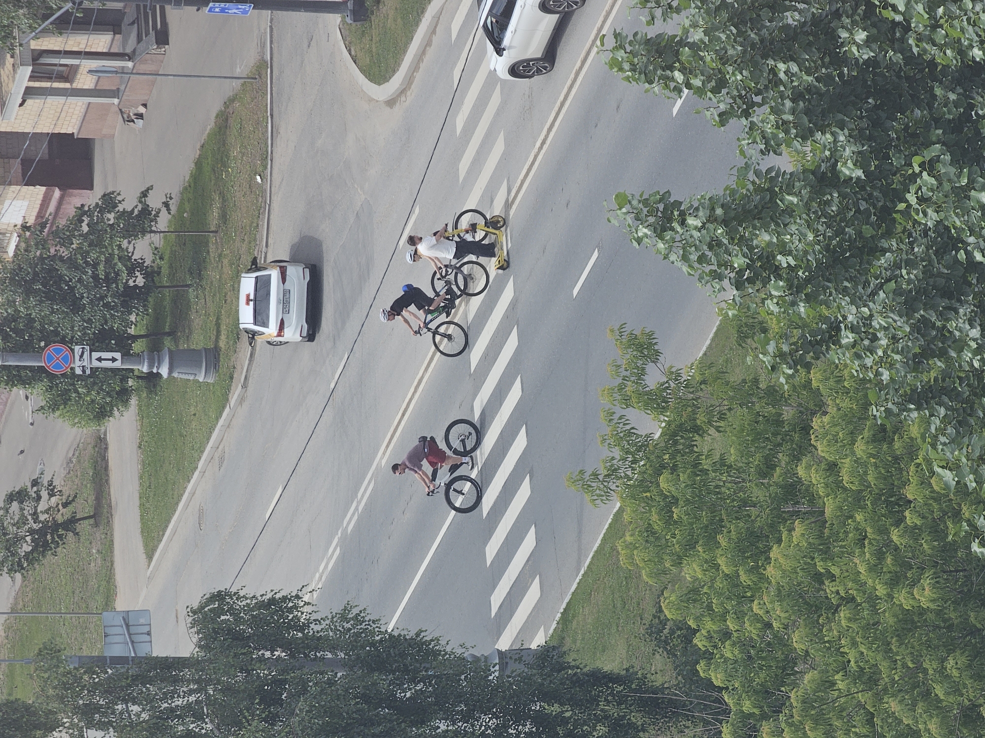 An aerial, eye-level shot captures a busy street scene in Moscow, Russia. The image features a pedestrian crossing with white stripes demarcating it on a grey asphalt road. Three individuals are crossing the street, two on bicycles and one on a yellow electric scooter. The person on the left is a male cyclist with short hair, wearing a maroon shirt and red shorts, riding a large-wheeled bicycle. In the middle, another male cyclist, bald, in a black t-shirt and black shorts, rides a bicycle. To the right of him, a man wearing a white t-shirt and dark pants stands on a yellow electric scooter, holding onto the handlebars. A white SUV is visible on the right side of the pedestrian crossing, and a white car is stopped on the road in the background. Trees with green foliage line the sides of the road, and a sidewalk is visible beyond the trees. The overall lighting suggests it is daytime.