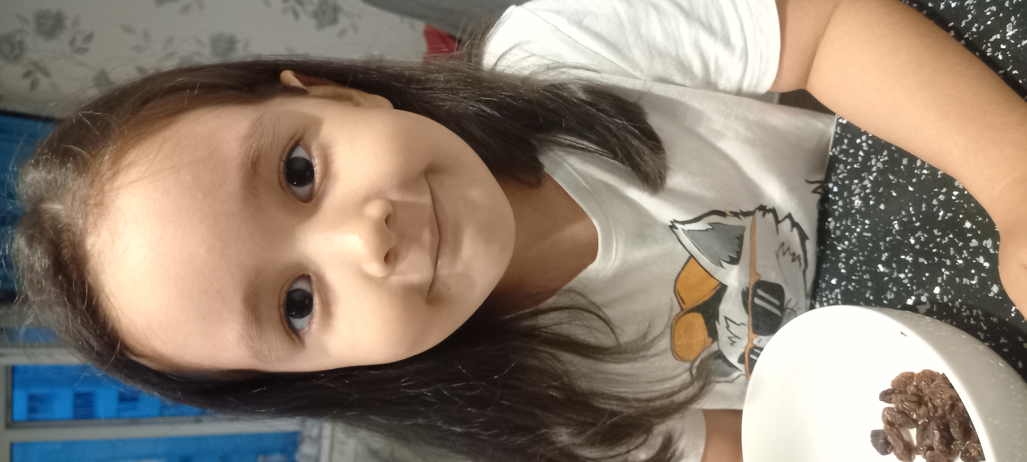 A young girl with dark hair and brown eyes is looking at the camera and smiling. She is wearing a white t-shirt with a graphic print of a raccoon wearing sunglasses. In front of her is a white bowl containing raisins. She is sitting at a table with a black speckled surface. The background is blurred but appears to be an indoor setting with a window showing a blue building. The overall lighting suggests an indoor environment. There are no discernible markers to indicate the city of Novosibirsk, Russia.