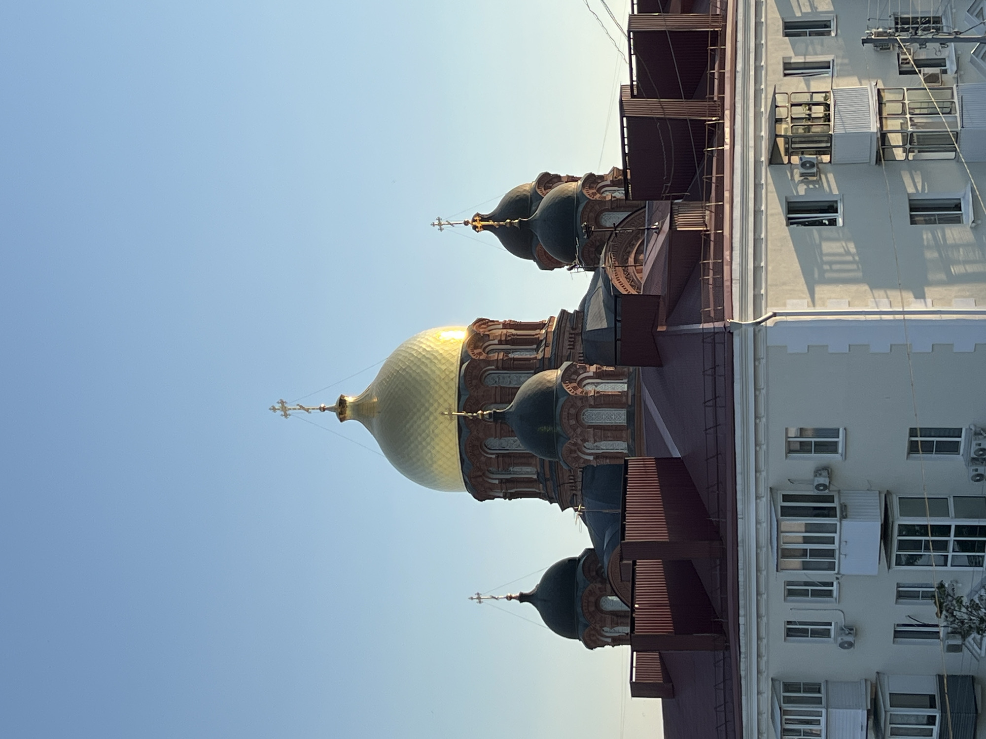 The image depicts a close-up view of an Orthodox church with multiple domes, some gilded and others dark green, set against a clear blue sky. The church's architecture features intricate detailing with arched windows and decorative elements. Adjacent to the church is a multi-story building with a light-colored facade. This building has numerous windows, some of which are reflective and appear to be air-conditioned. The scene is bathed in sunlight, suggesting it is daytime, likely late afternoon or early morning due to the warm glow on the gilded dome. The composition is vertical, with the church occupying the left side of the frame and the adjacent building on the right. There is no visible text or any people in the frame. The overall impression is one of architectural grandeur and a juxtaposition of religious and modern urban structures.