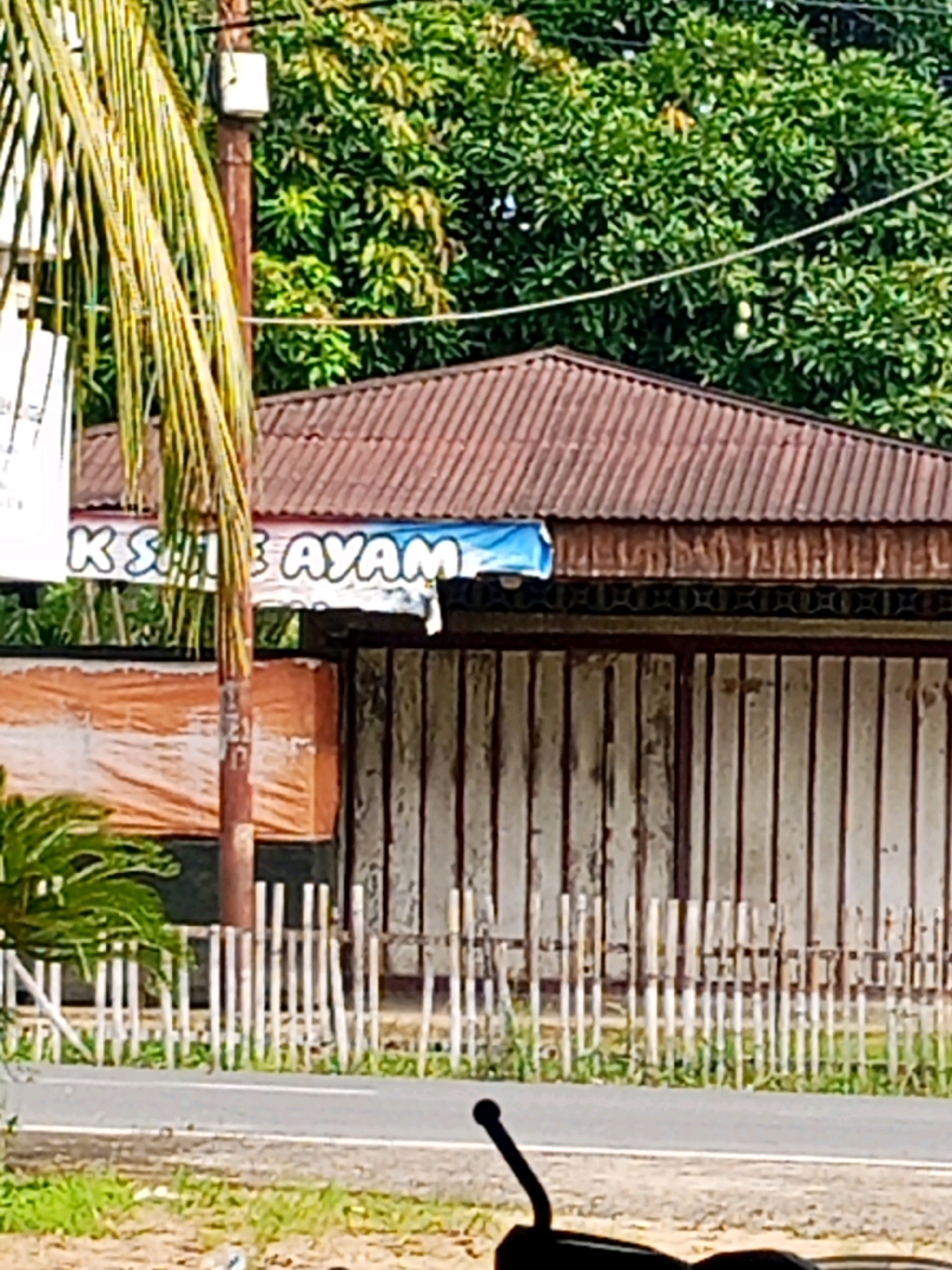 A building with a corrugated metal roof and vertical white wooden doors stands behind a white picket fence. A portion of a blue banner with white lettering is visible on the front of the building. A street runs in front of the fence, with a motorcyclist's handlebars visible in the foreground. Lush green trees and vegetation surround the building. The scene is set in Jinang Kampung Pansur, Indonesia.