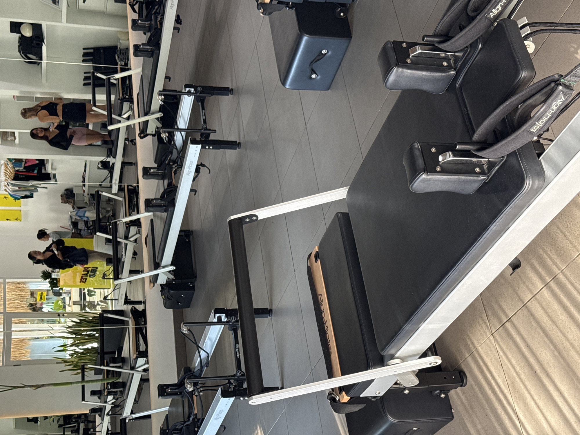 Here is a detailed description of the image:
The image depicts a pilates studio or fitness center with multiple pilates machines arranged in rows. The studio appears to be in a modern, well-lit space with a neutral color palette. Large windows provide natural light and views of the outside vegetation.
Several people are visible in the background, some of whom appear to be exercising or engaged in activities related to pilates or fitness. The pilates machines are the main focus, with one in the foreground positioned prominently in the frame. The machines are black and white, with the brand name "Align-Pilates" visible on the frame.
The environment includes elements of urban design, such as the tiled floor and structured layout of the machines. The presence of plants near the windows adds a touch of nature to the space.
Based on the signs and the style of the studio, it is likely located in the city of Ubud District, Indonesia.
