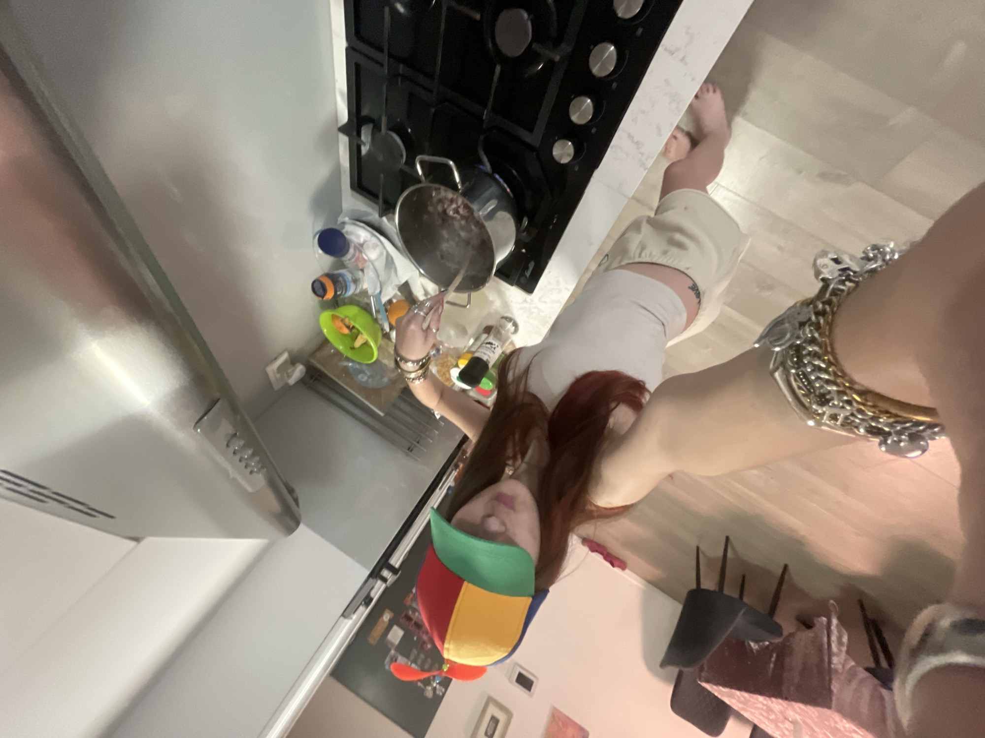 A woman wearing a multi-colored propeller hat and jewelry on her wrist and arm is cooking at a gas stove. She is stirring a pot of beans. The location appears to be a kitchen in Raanana, Israel, based on the presence of a gas stove and the general appearance of the interior. A green bowl, bottles, and other cooking items are near the stove. A dining area with chairs is visible in the background.