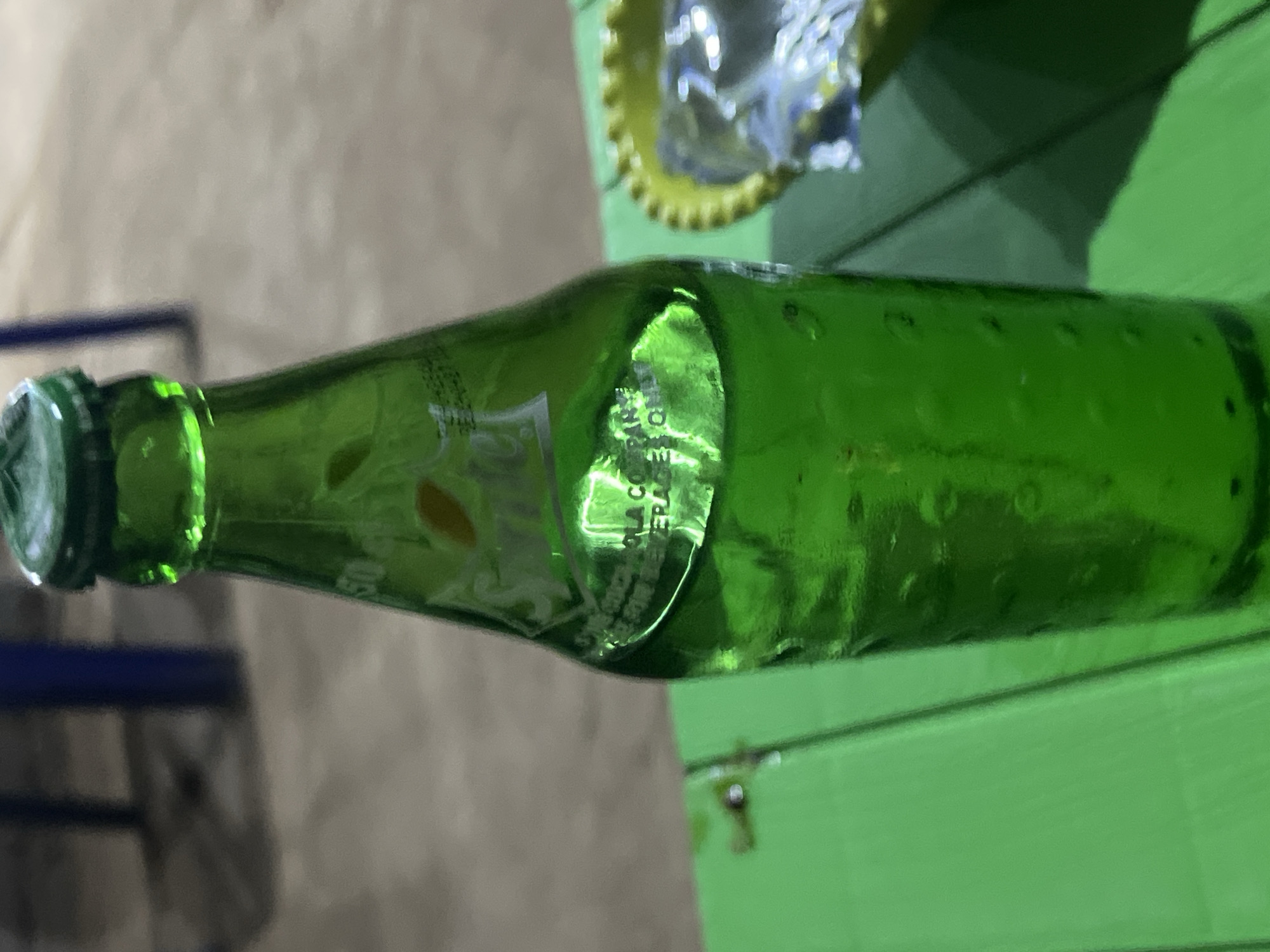 A green glass Sprite bottle is positioned horizontally on a bright green, possibly wooden, surface. To the left of the bottle, a blurred blue metal chair is partially visible against a beige concrete background. To the right of the bottle, a crumpled piece of foil in a yellow wrapper sits atop the green surface. Minute details, such as condensation droplets on the bottle's exterior, are discernible. Location: Possibly Jalingo, Nigeria.