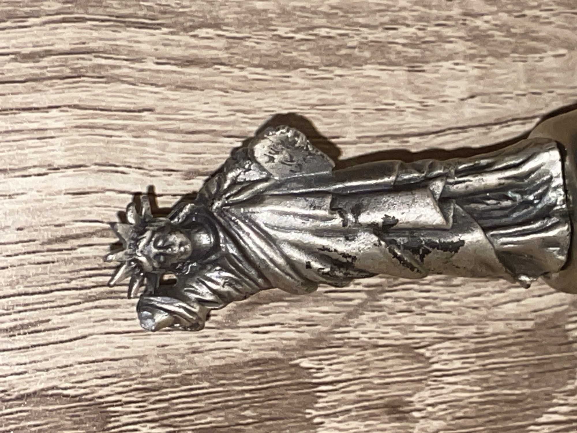 A miniature silver-colored statue of the Statue of Liberty stands on a wooden surface. The statue is detailed with a crown, a robe, and a tablet. The wooden surface has a vertical grain pattern.