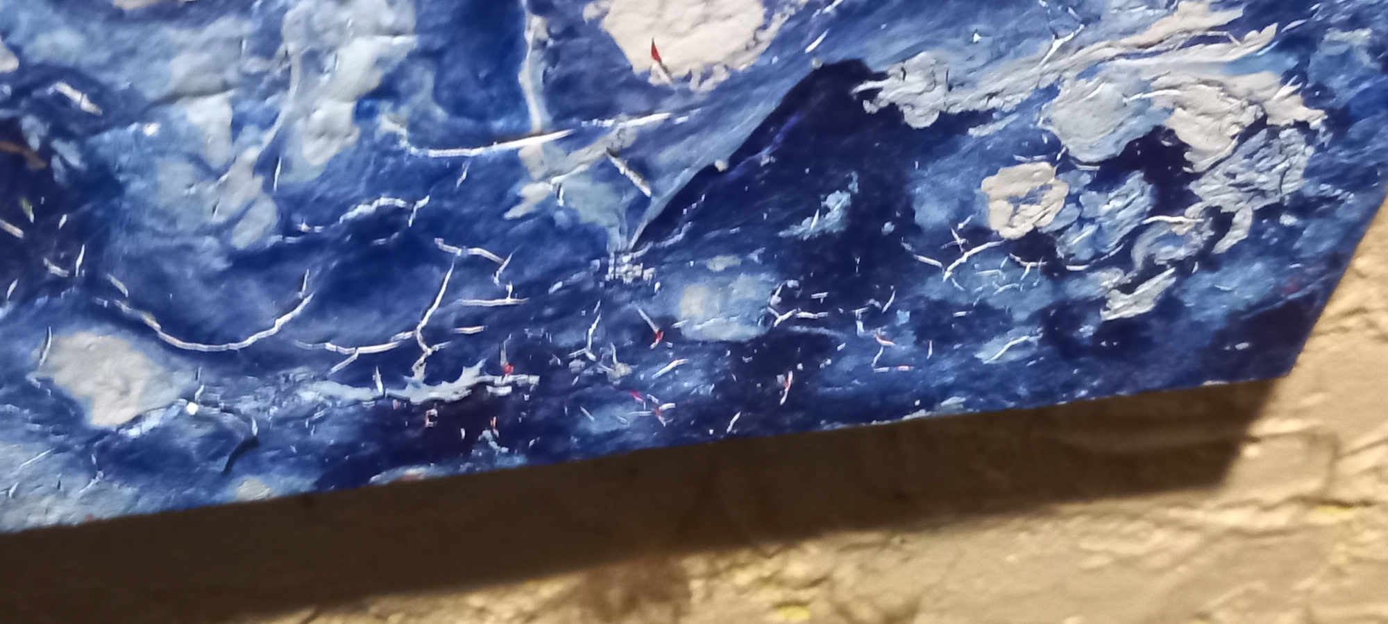 This image depicts a fragment of a highly textured, abstract surface, predominantly deep blue, white, and light gray, presented as a scene situated in Kryvyi Rih, Ukraine. The central subject is a thick, irregular blue material, possibly an artwork or decorative element, characterized by a complex pattern of white and light gray raised areas and fine, intricate crack-like lines. These white and gray elements create a highly tactile and distressed appearance across the varying shades of blue. Several small, bright red flecks are interspersed within the blue and white textures, particularly visible in the lower-left section of the blue object. The blue surface is angled, occupying the majority of the right and center of the frame. To its left, a vertical section of a lighter, uniformly textured, light brown or beige surface is visible, likely a wall. This beige surface has a rough, stucco-like finish. The blue object casts a distinct shadow onto the beige surface, indicating a light source from the upper-left. No human figures, specific actions, or other discernible objects beyond these textured surfaces are present.