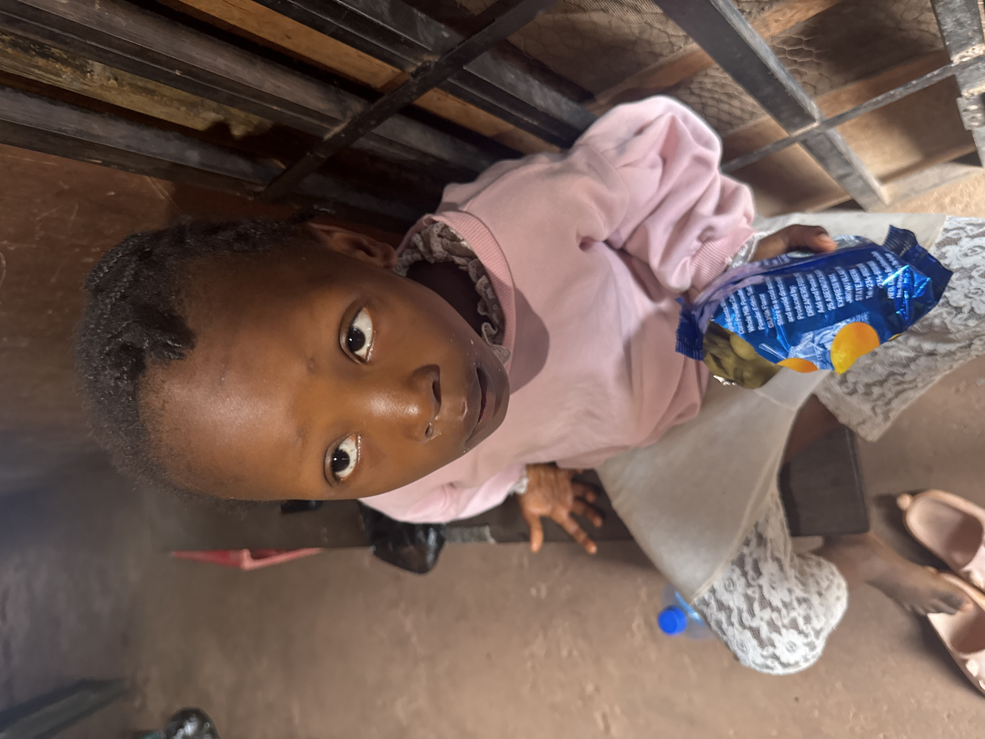 The image depicts a young Black child, likely female, seated and looking upwards. The child is wearing a pink sweatshirt with a decorative lace collar and light-colored pants with a lace pattern. They are holding a blue and orange snack bag in their right hand. A blue water bottle is visible to the left of the child. The background appears to be an indoor setting with a dirt floor and a wooden structure with metal bars. The overall lighting is dim. While the image provides a glimpse into the child's immediate surroundings, specific details placing it within the city of Enugu, Nigeria, are not directly visible.