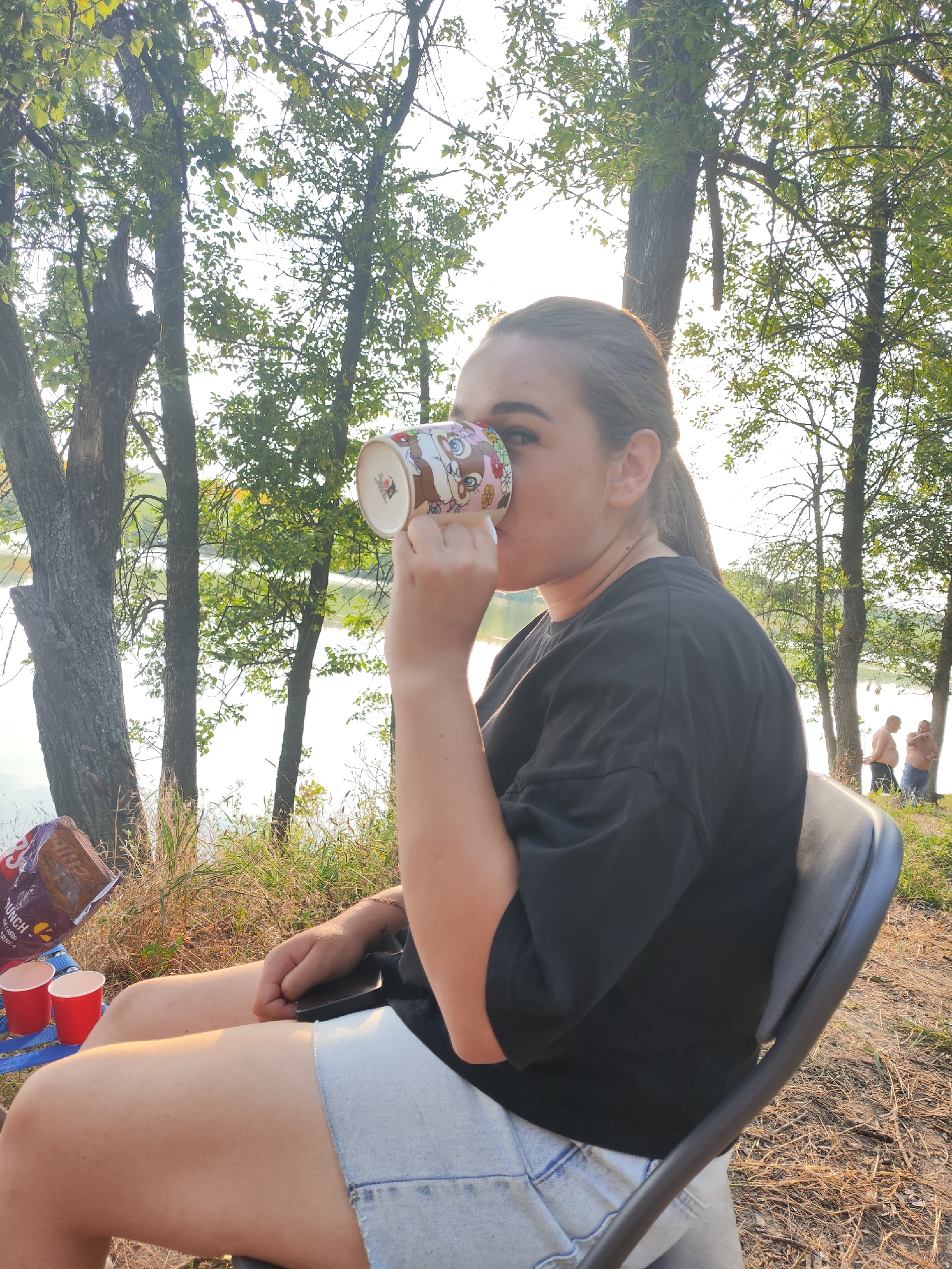 A young woman with her hair in a ponytail, wearing a black t-shirt and denim shorts, sits in a black chair outdoors and drinks from a mug. In front of her, on a blue surface, are two red cups and a bag of snacks. Behind her, a body of water is visible, with trees and two shirtless men in the background. The scene appears to be in Găleștii Noi, Moldova, during daylight.